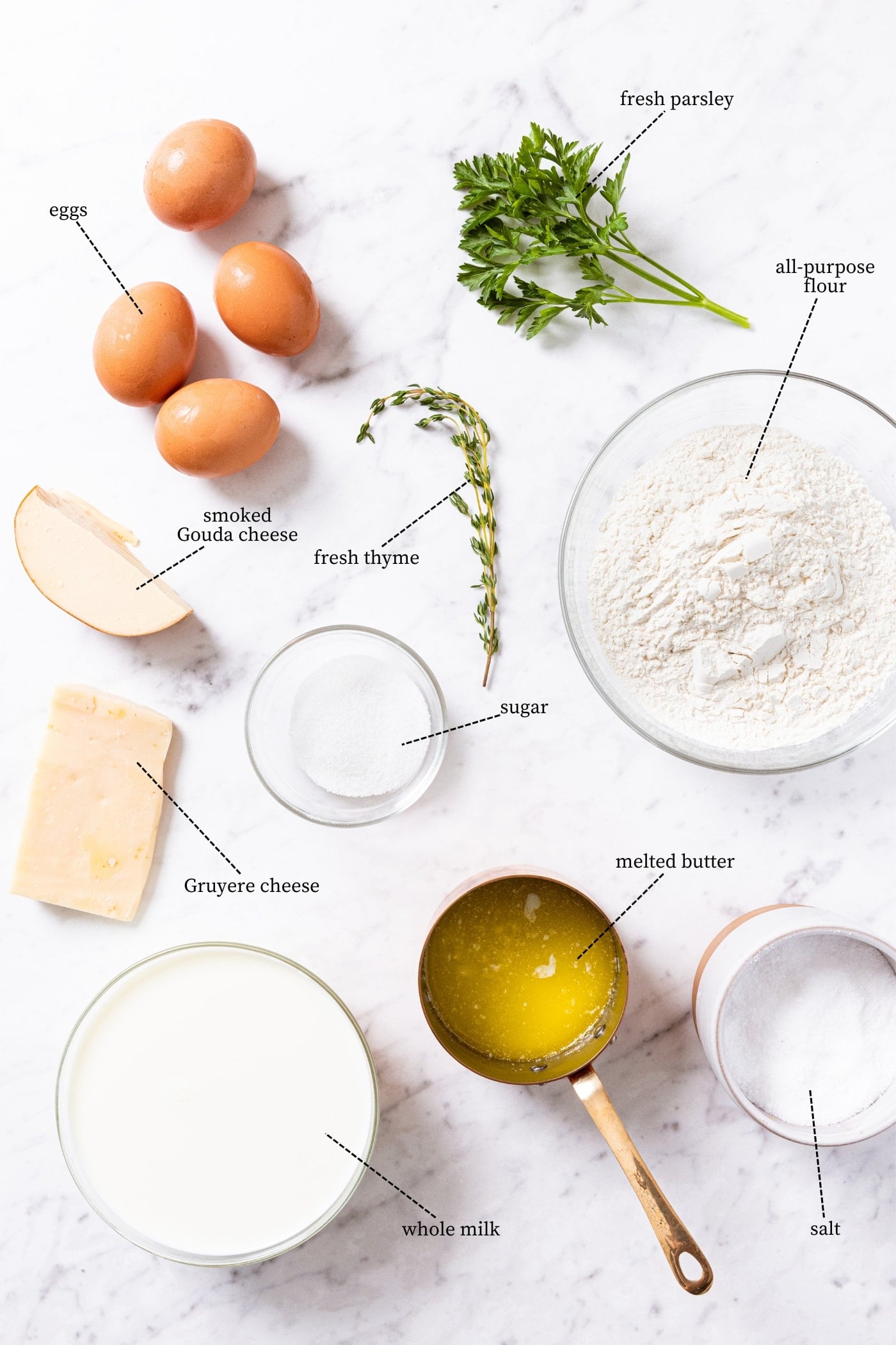 Marble surface filled with all ingredients needed to make cheese popovers including eggs, flour, herbs, cheese, butter, and more.