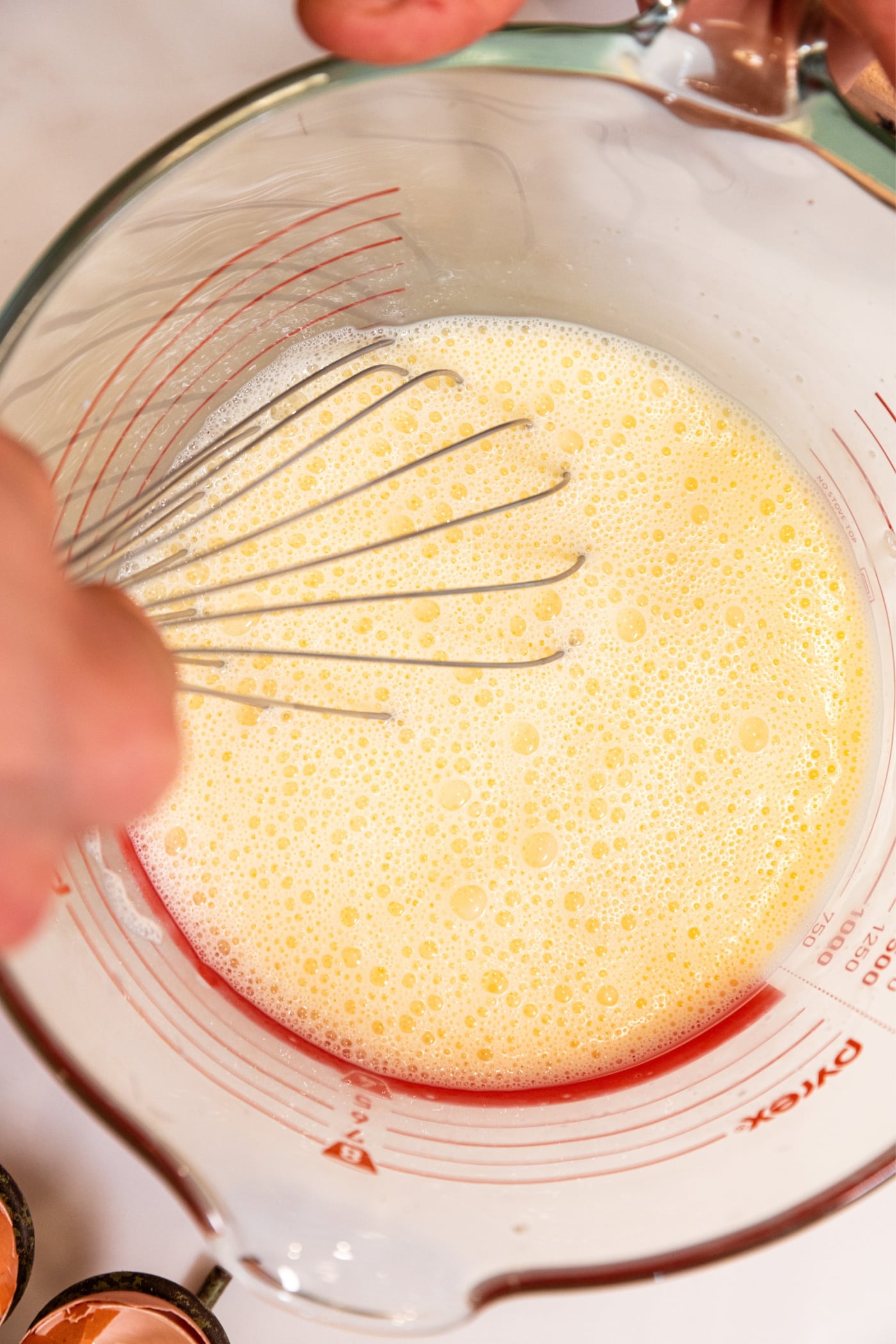 Hand holding metal whisk mixing together eggs and milk in a large glass measuring cup on white countertop.