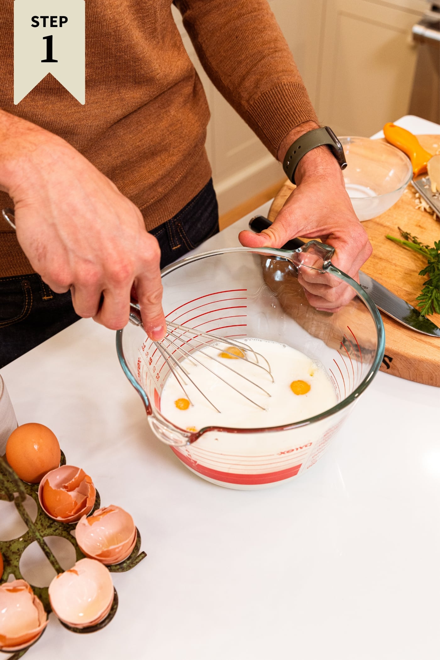 Two hands mixing eggs and milk mixture in glass measuring cup with egg shells to the side all on white countertop.