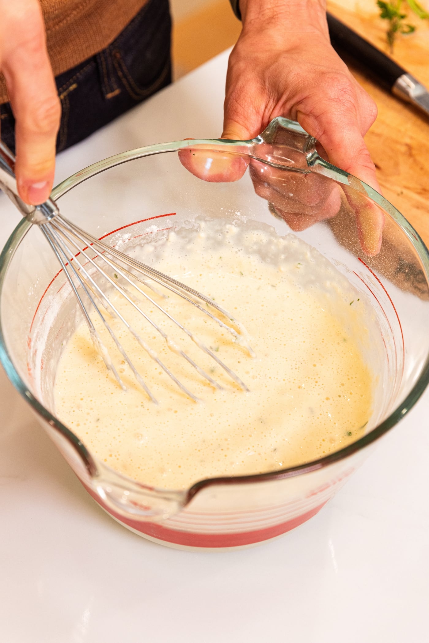 Hand holding whisk mixing together all ingredients in a glass measuring cup all on a white surface with wood cutting board in background.
