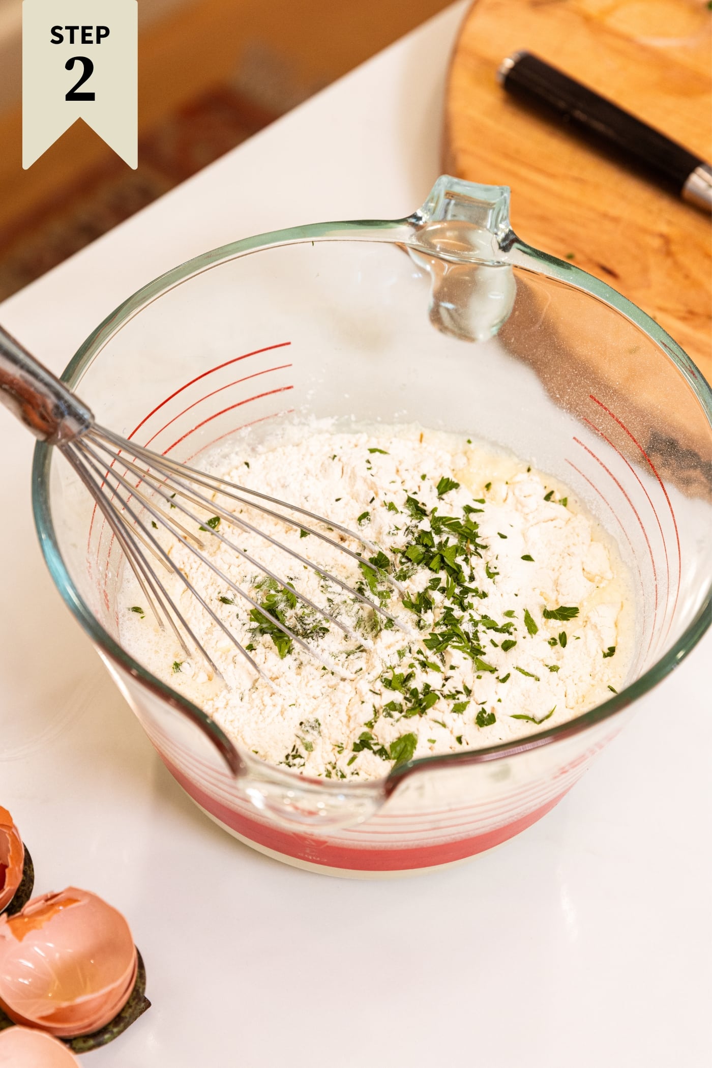 Glass measuring bowl filled with egg and milk mixture with flour and chopped herbs resting on top all on white countertop.