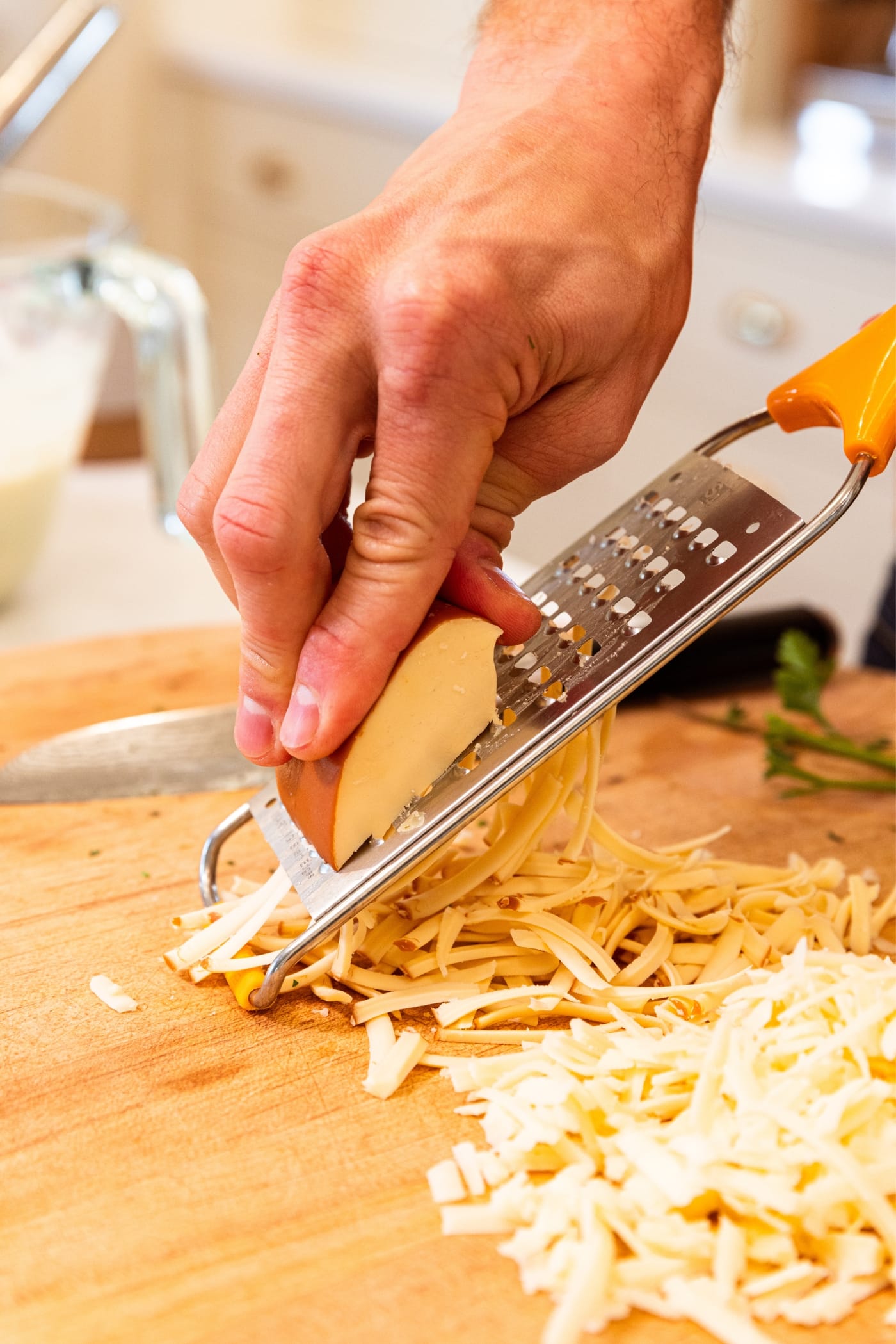 Hand cutting cheese into strip on cheese grater with pile of cheese sitting to the side on wood cutting board.