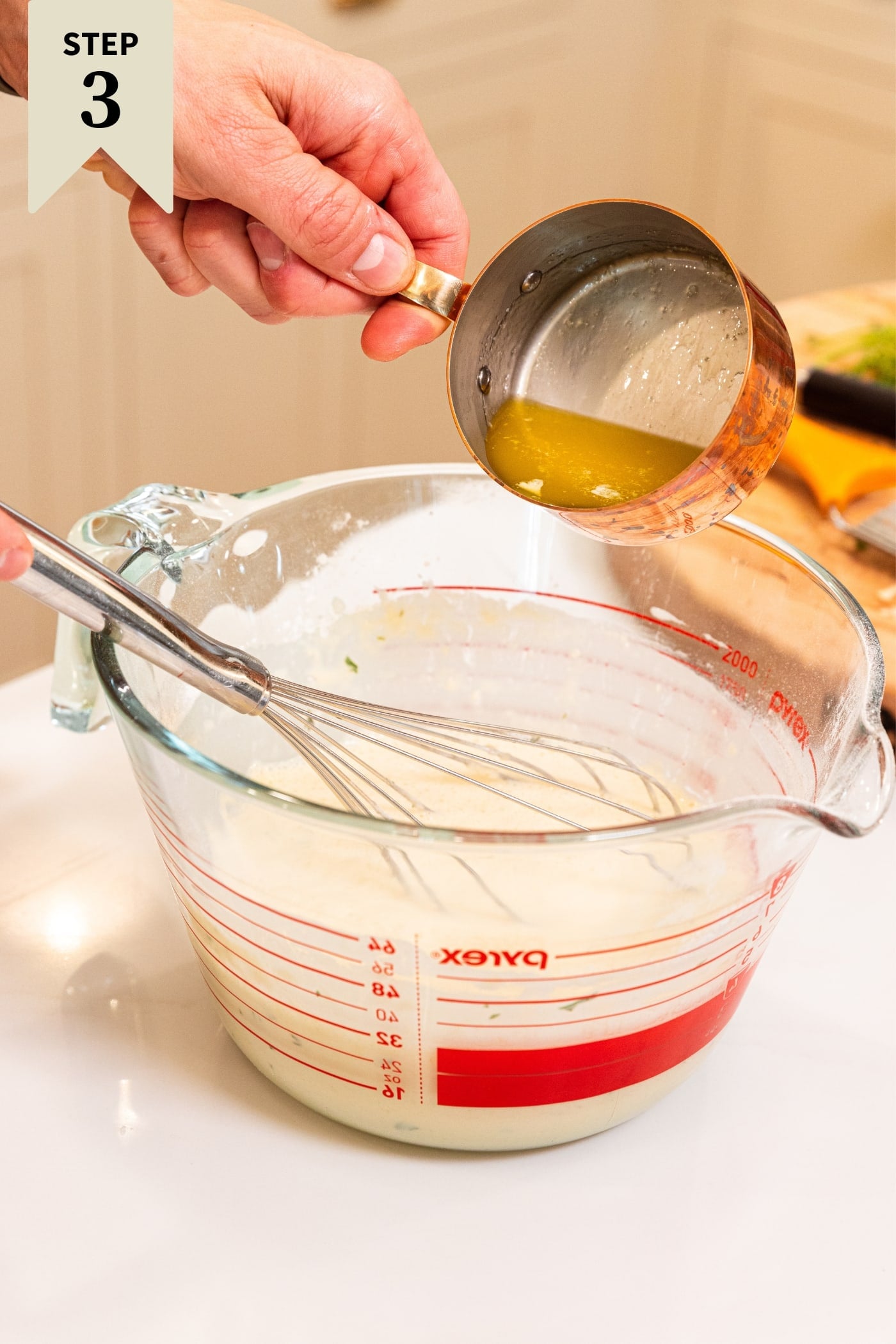 Hand holding small copper pot with melted butter in it about to be poured into glass mixing bowl filled with milk and flour mixture all on white countertop.
