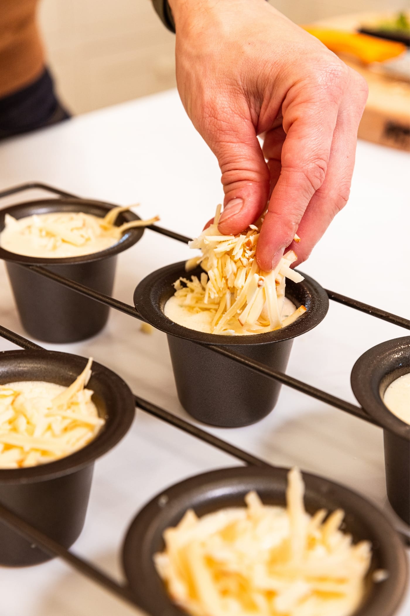 Hand placing pieces of shredded cheese on top of a popover in popover pan with more popovers in background without cheese on them all on white surface.