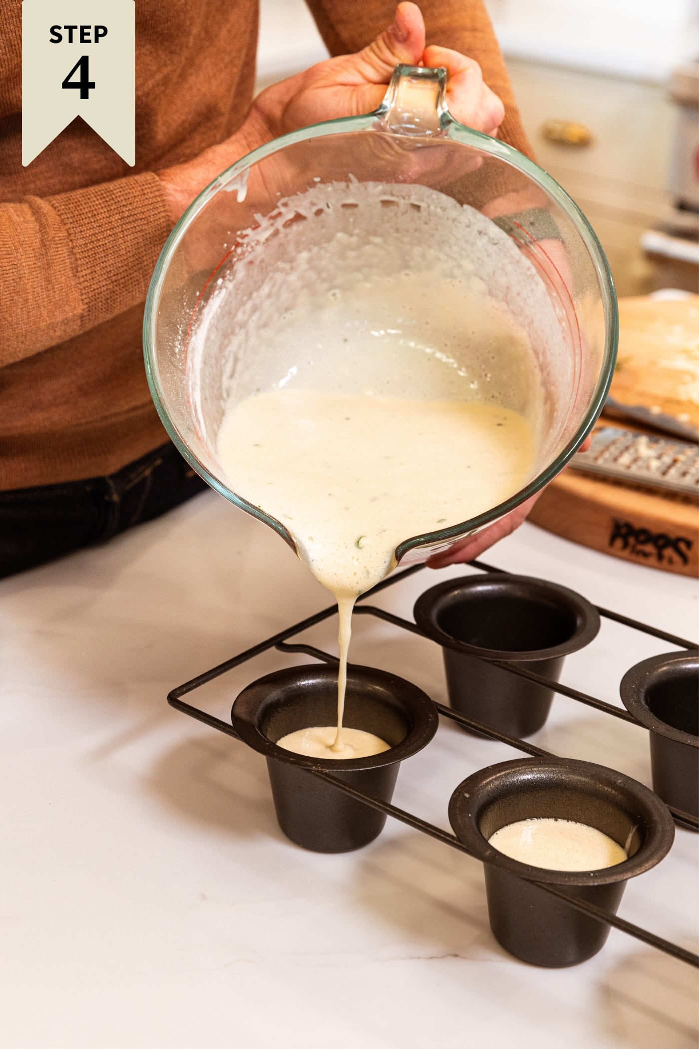 Hand holding glass measuring cup filled with milk and flour mixture pouring into popover pan on white countertop.
