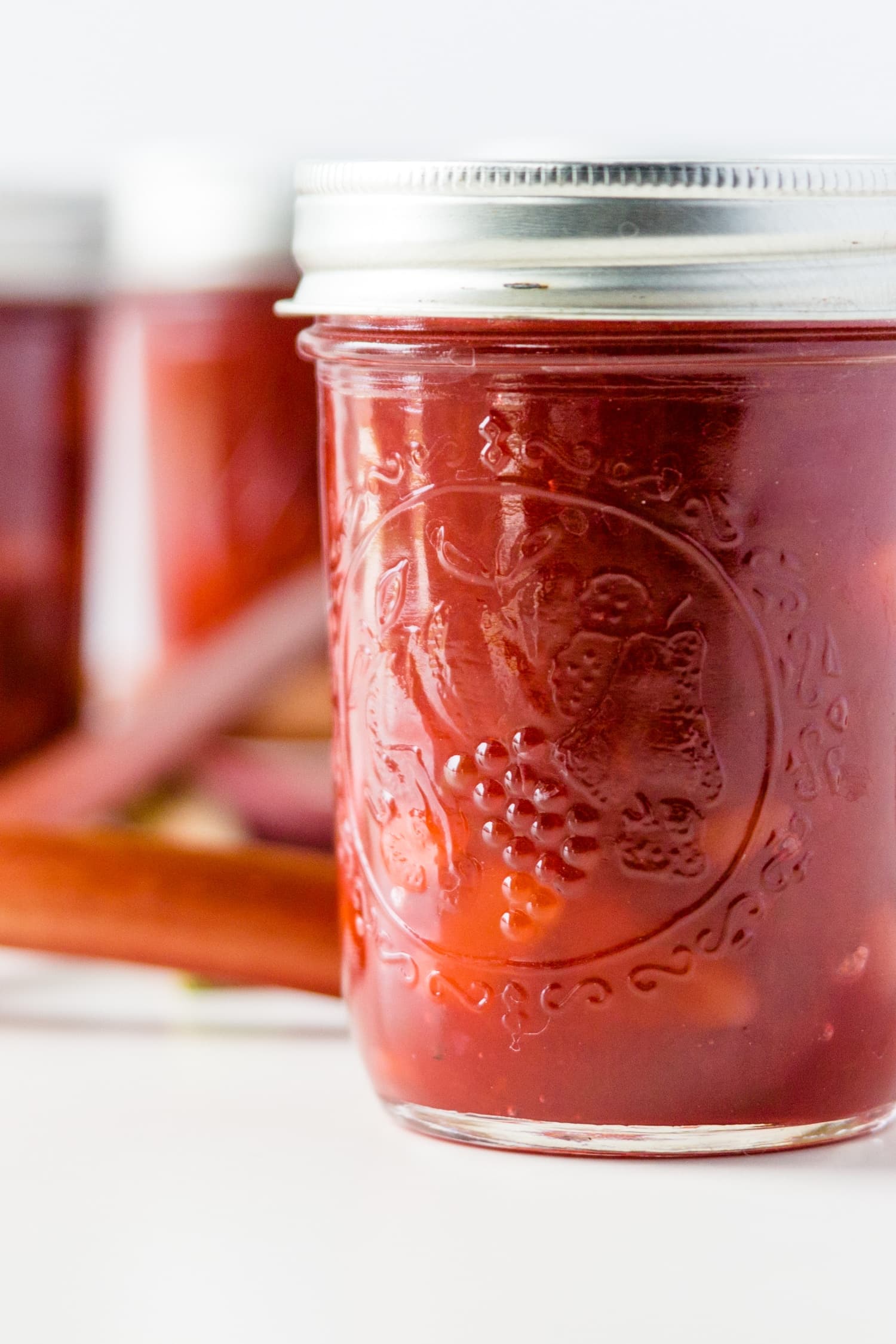 Jar of rhubarb jam with extra jars and rhubarb stalks in background on a white surface