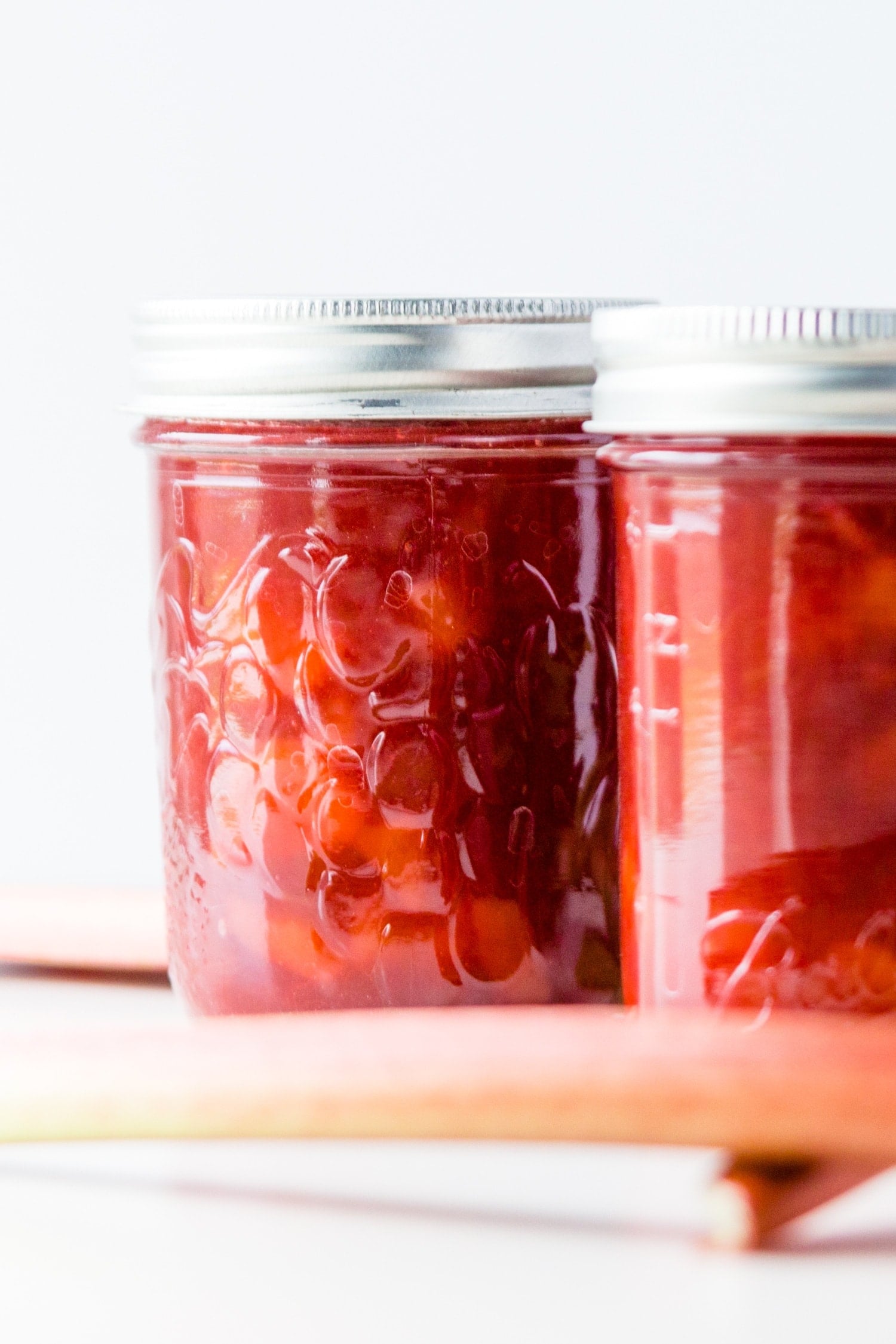 Jar of rhubarb jam with extra jars and rhubarb stalks on a white surface