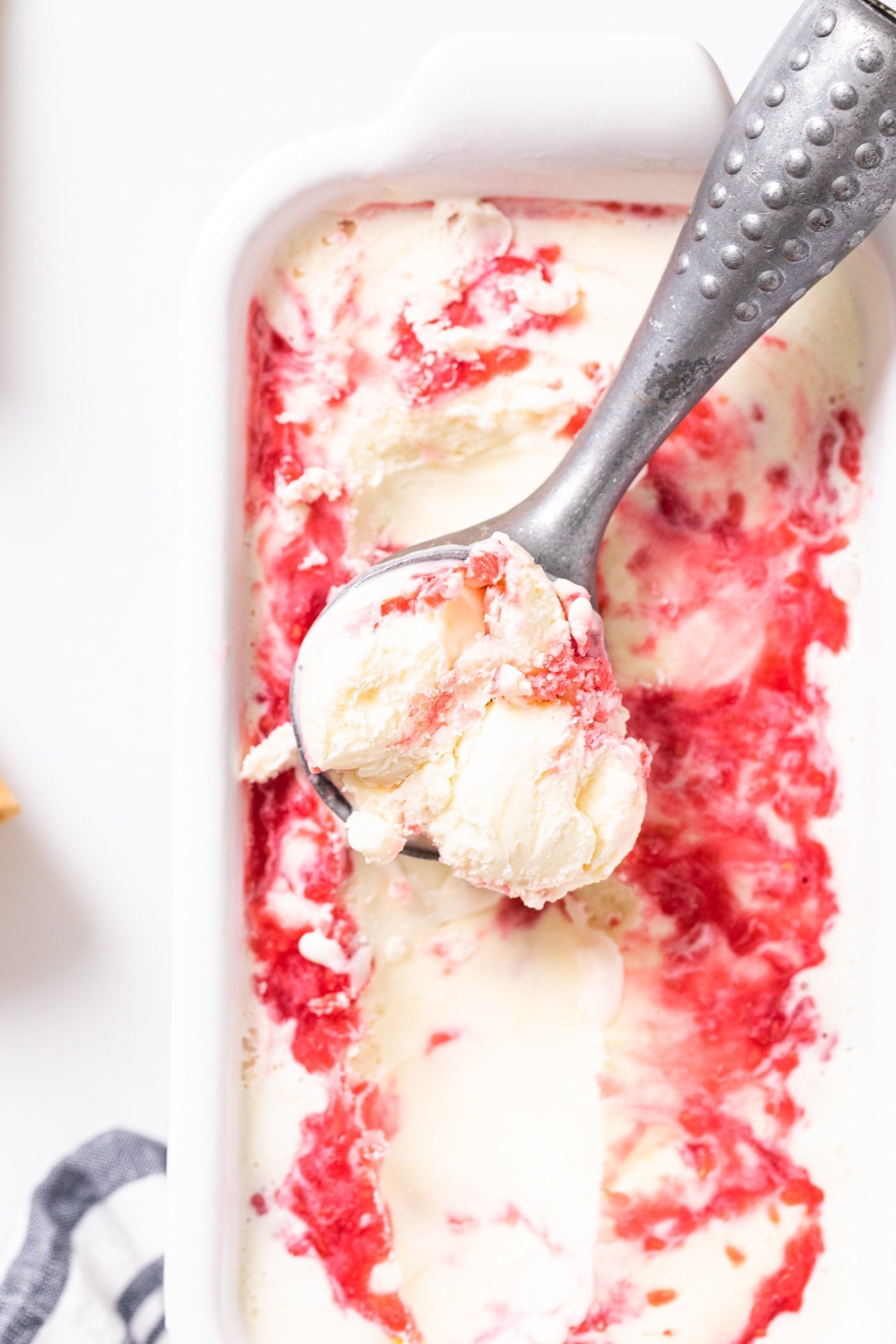Top down view of white container with silver scoop filled with white and red colored ice cream sitting on white countertop