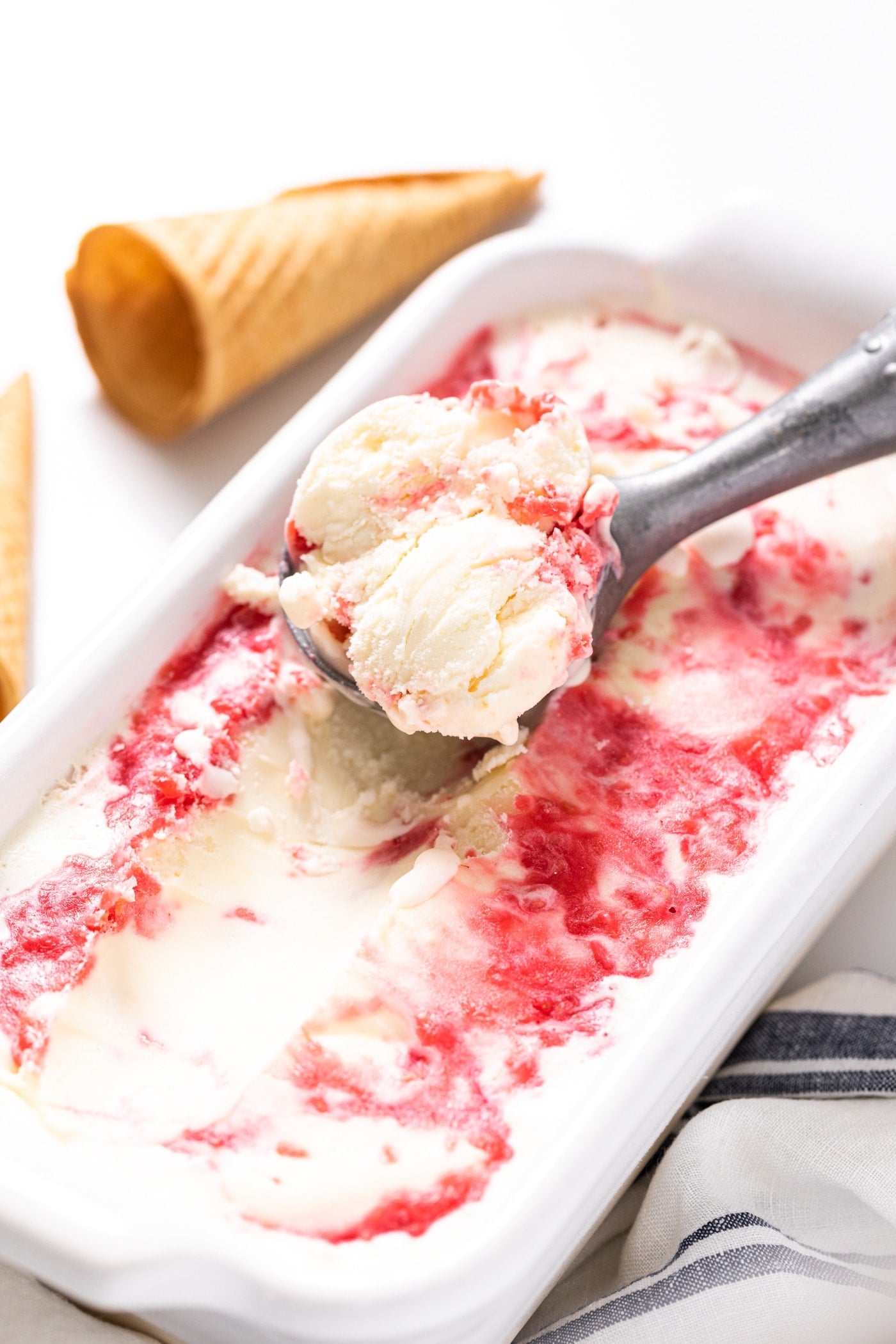 White loaf pan filled with white ice cream swirled with raspberry sauce with empty ice cream cones in the background all on white countertop