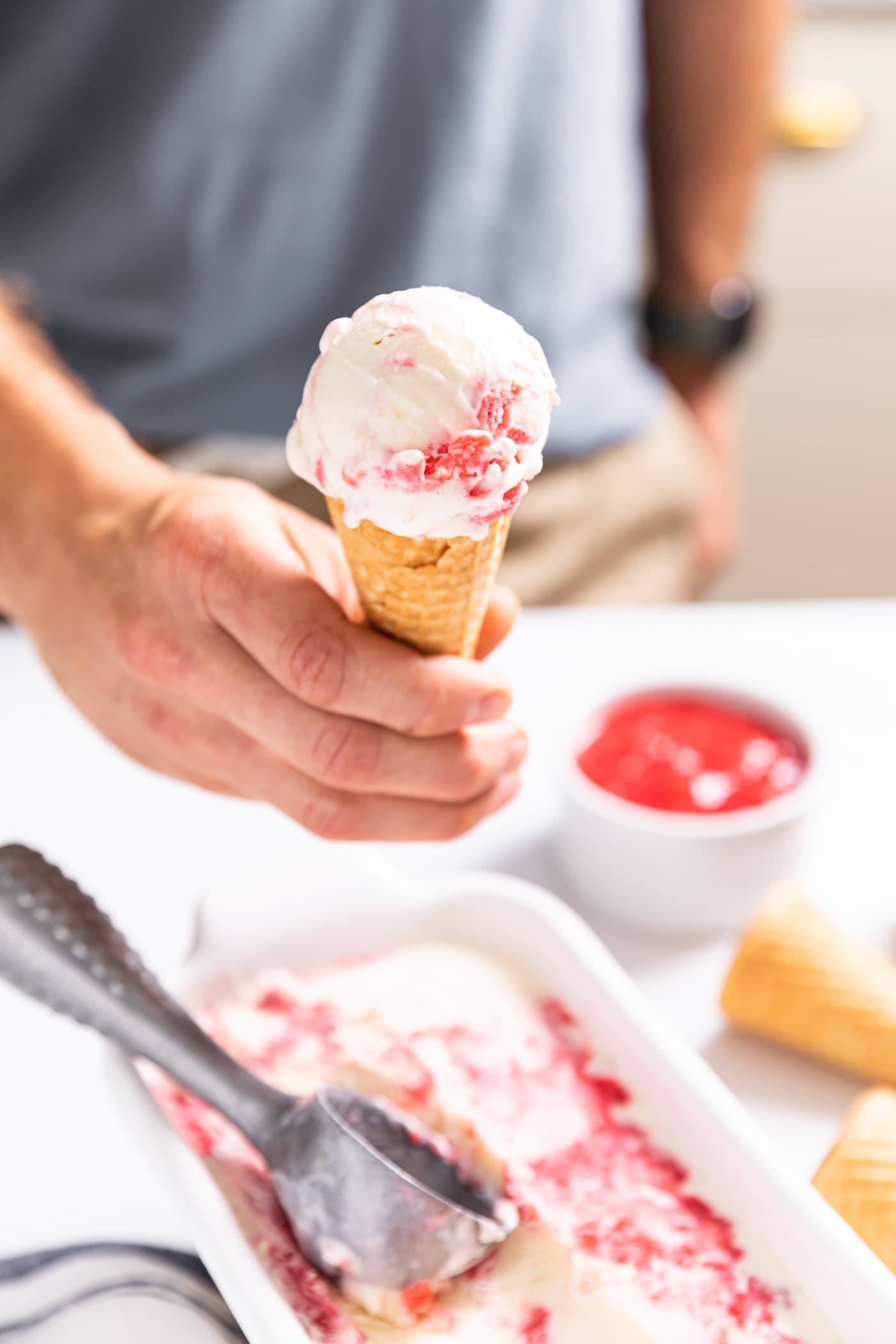 Hand holding ice cream cone filled with scoop of white and raspberry swirled ice cream with rest of container and ice cream scoop sitting on countertop below