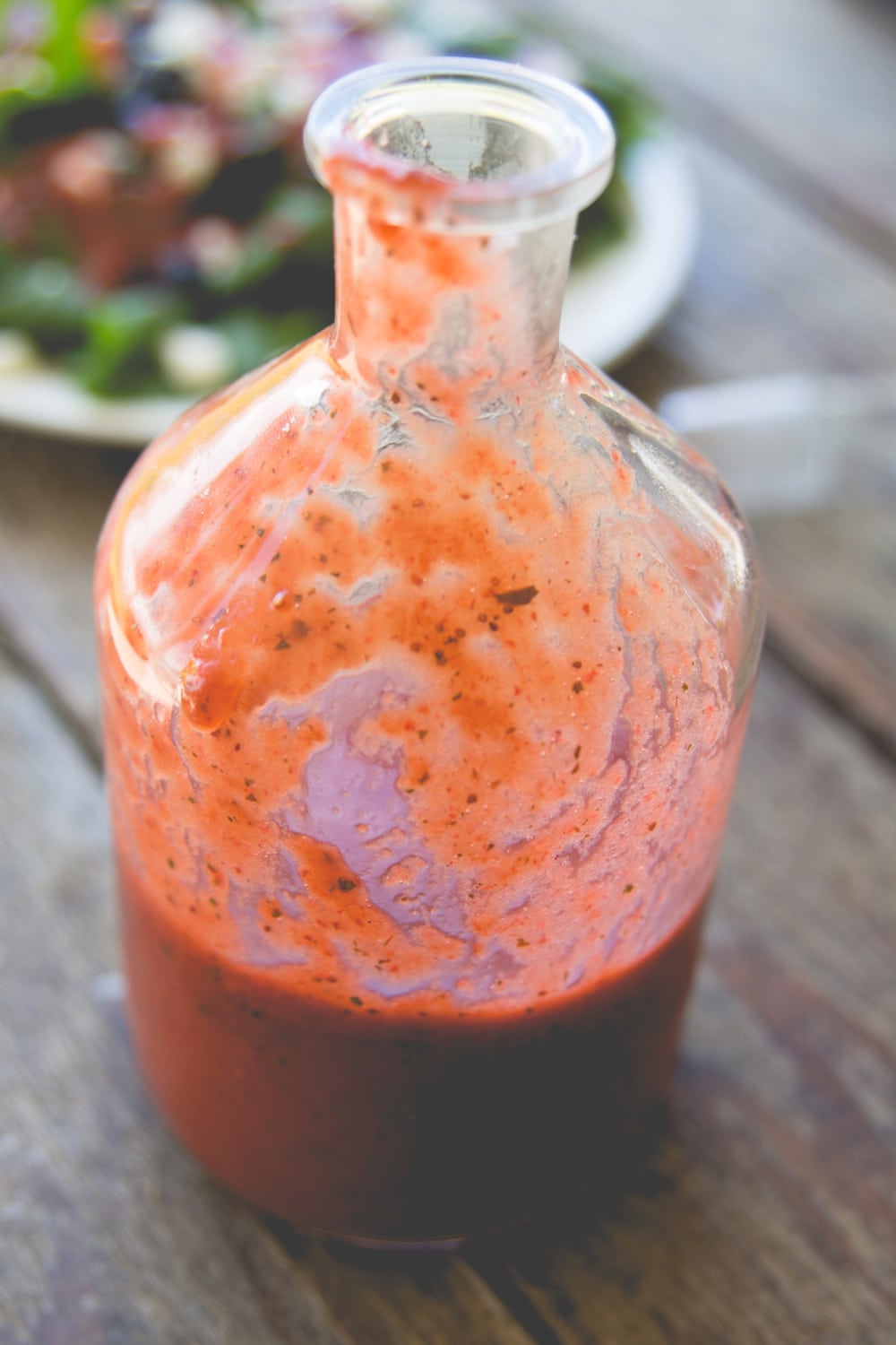 Glass container filled with pink colored strawberry vinaigrette dressing with salad in background
