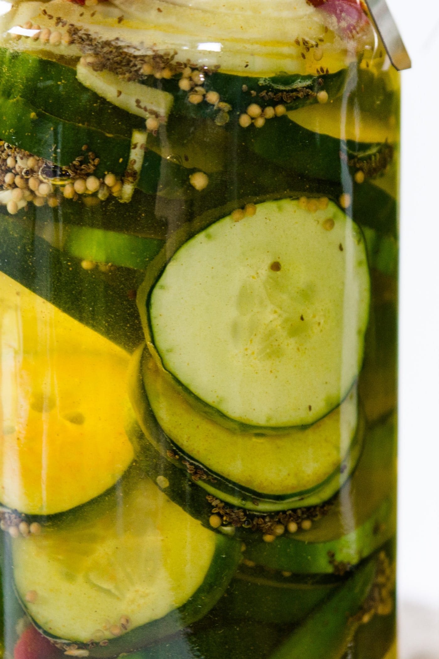 Glass jar of fridge pickles with cucumbers, onions, red peppers and celery seed in light yellow pickling liquid