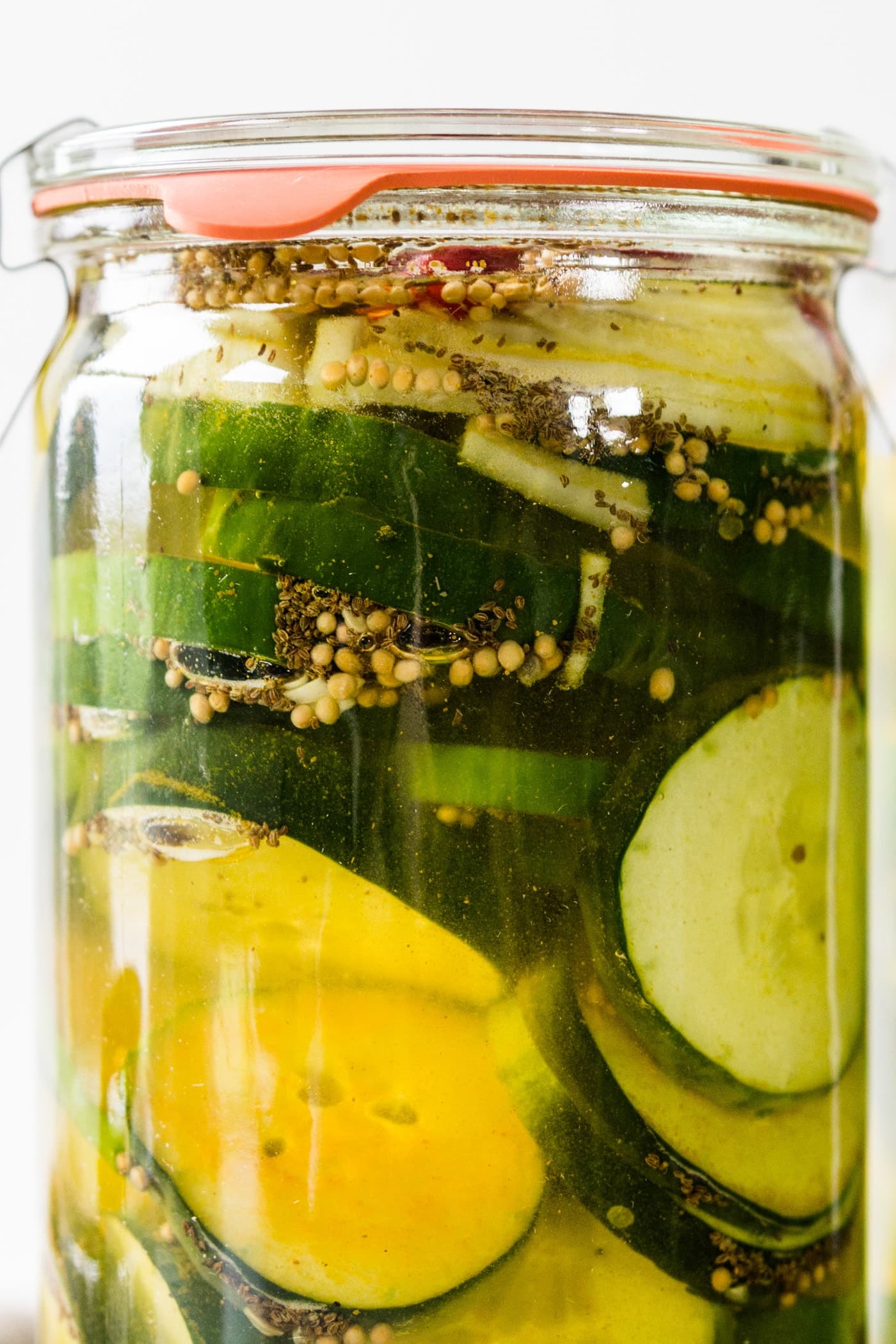 Glass jar of fridge pickles with cucumbers, onions, red peppers and celery seed in light yellow pickling liquid