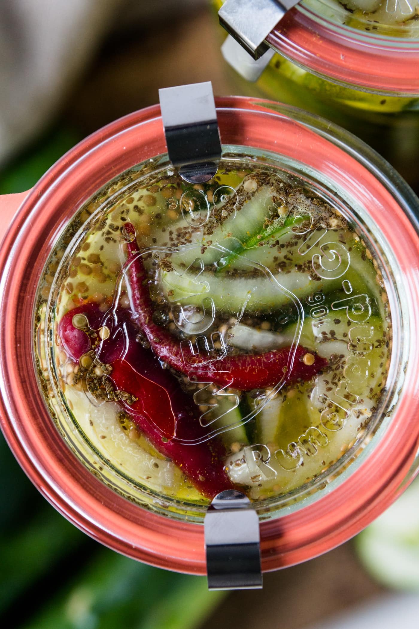 Top down view of Weck jar filled with fridge pickles with red peppers on top with extra cucumbers, cutting board and napkin in background