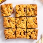 Top down view of nine peach crumb bars with golden top sitting on white countertop with sliced peaches around