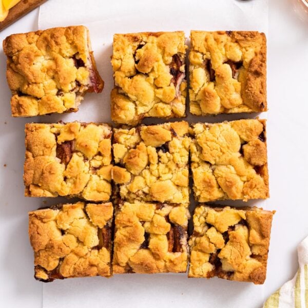Top down view of nine peach crumb bars with golden top sitting on white countertop with sliced peaches around
