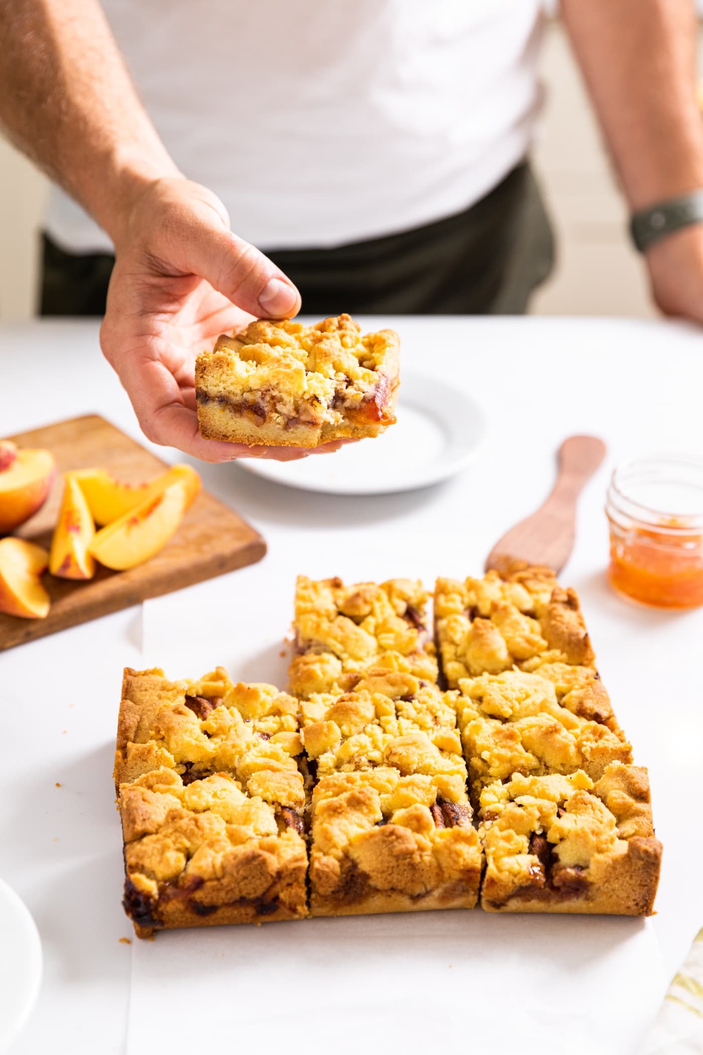 One hand holding single piece of a peach crumb bar with rest of bars sitting on white countertop below