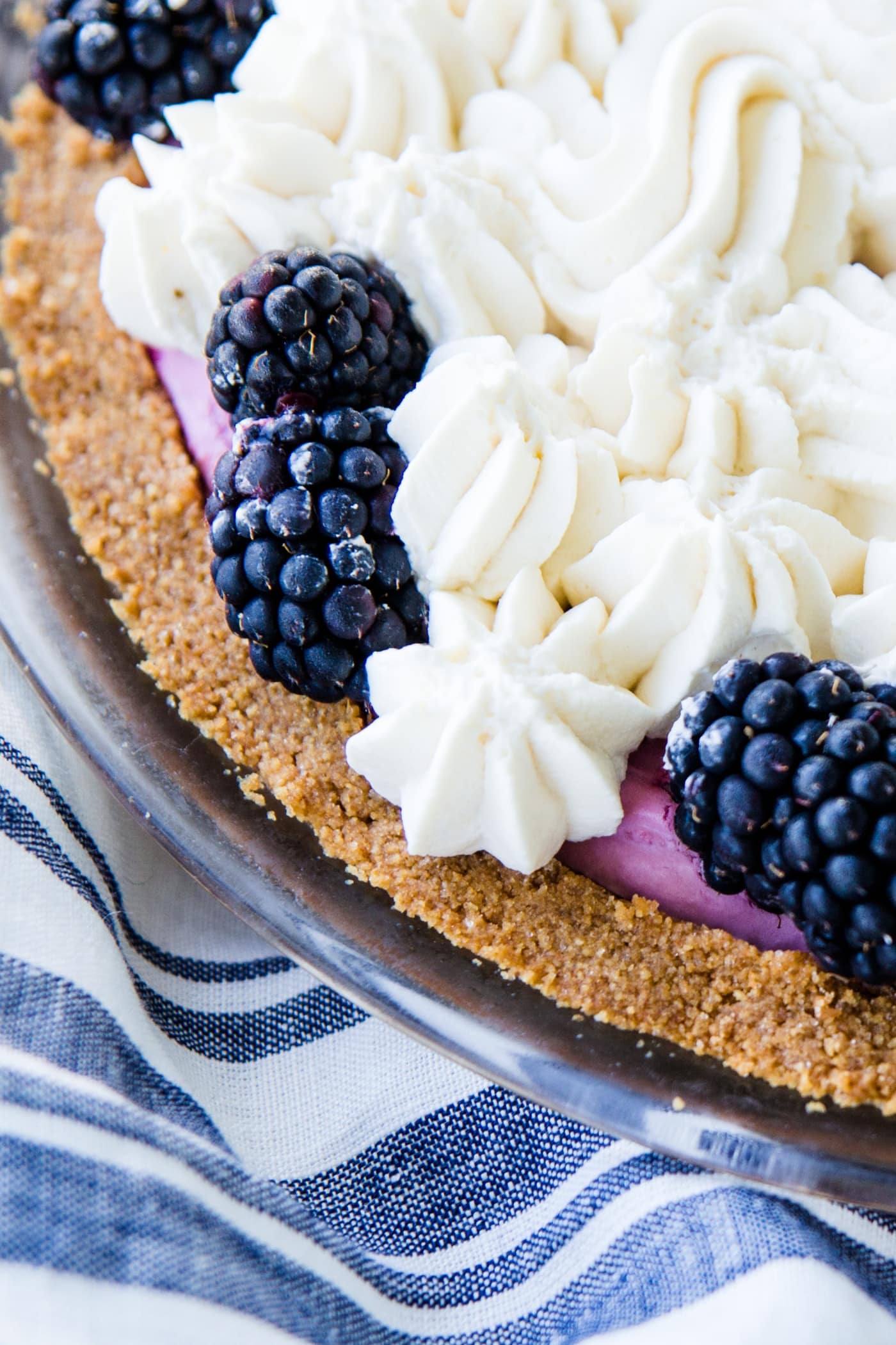 Top down view of edge of blackberry icebox pie sitting on a blue and white striped napkin with fresh whipped cream and whole blackberries on top