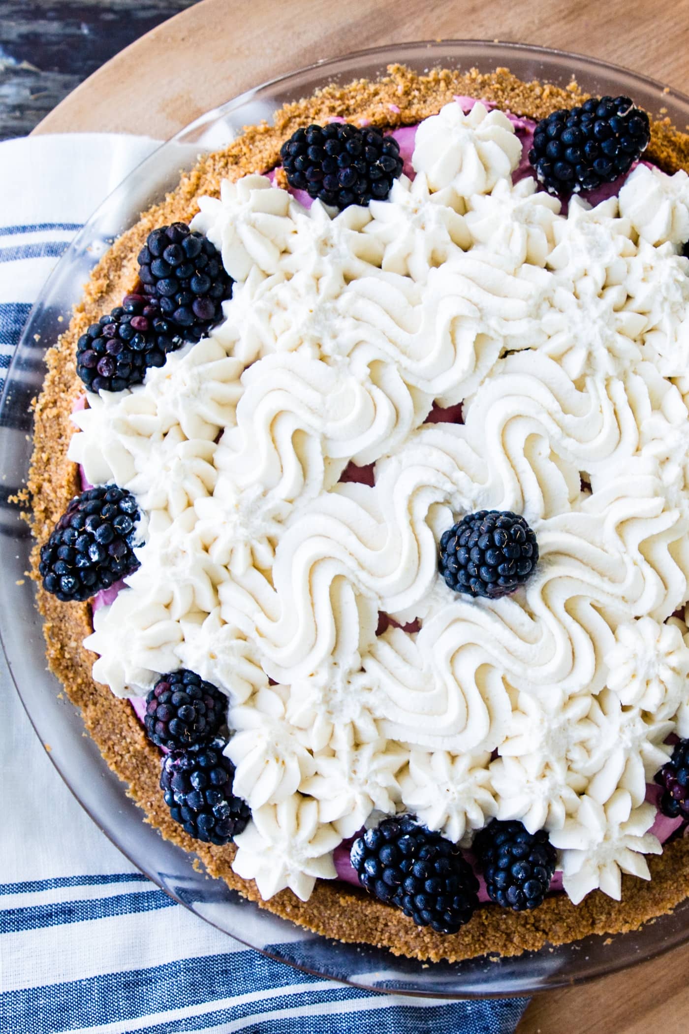 Top down view of blackberry icebox pie sitting on a blue and white striped napkin and wood serving platter with fresh whipped cream and whole blackberries on top