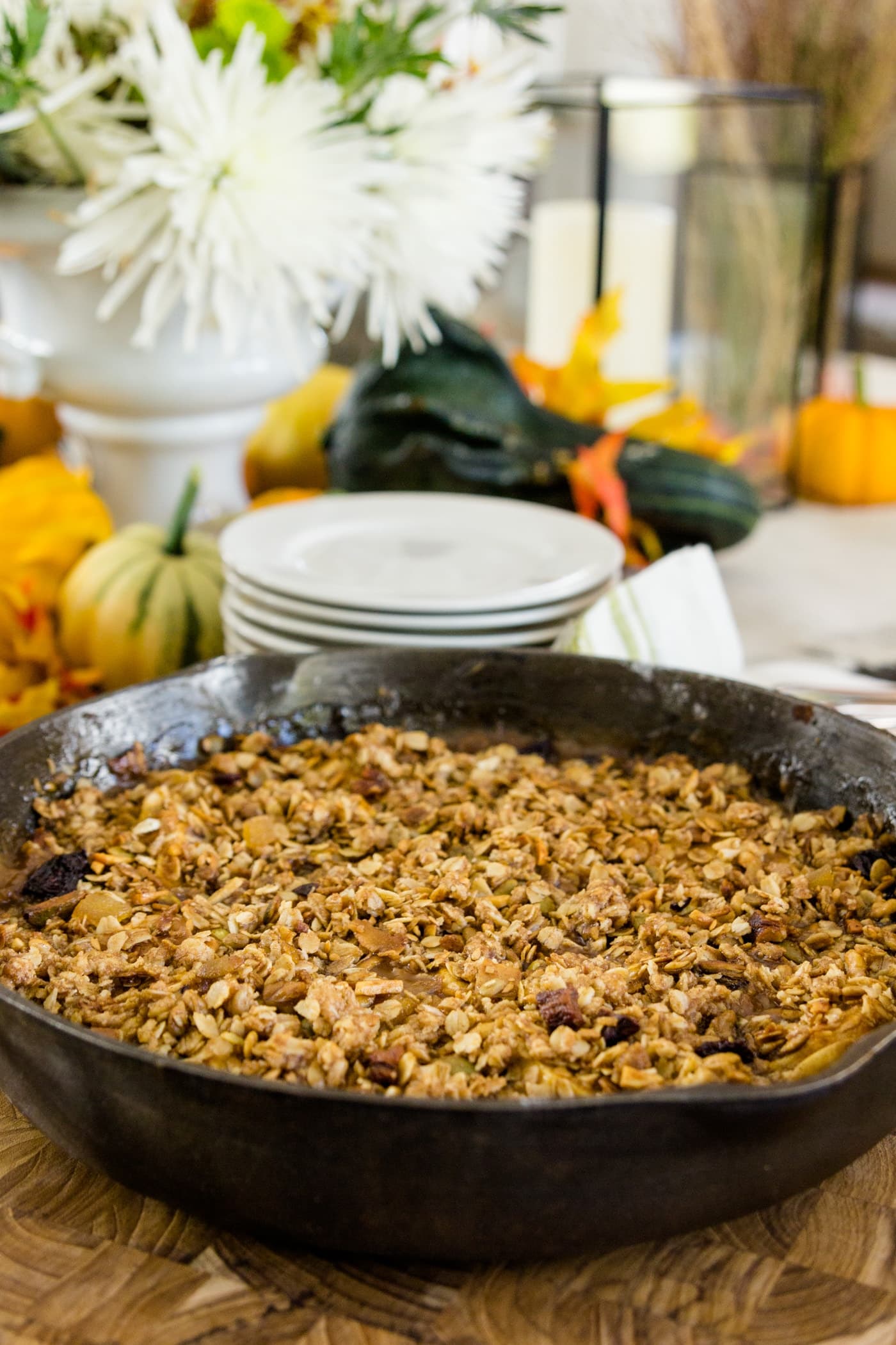 Skillet apple crisp sitting on wood cutting board with pumpkins, flowers, napkins and extra plates in background