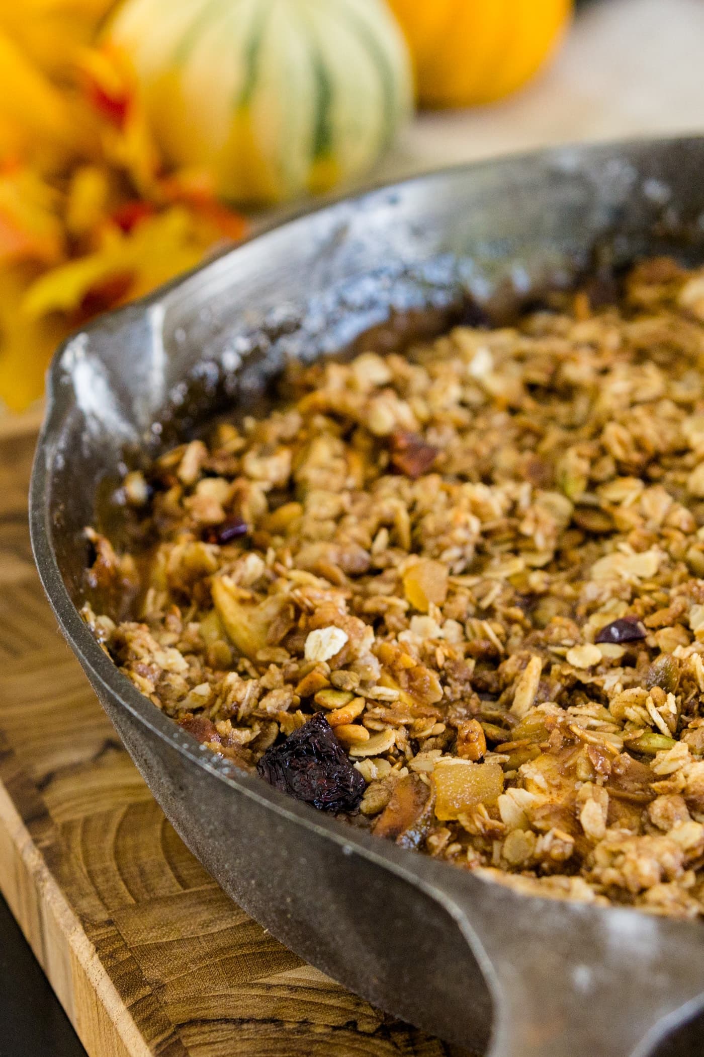 Skillet apple crisp sitting on wood cutting board with pumpkins and leaves in background on a black surface