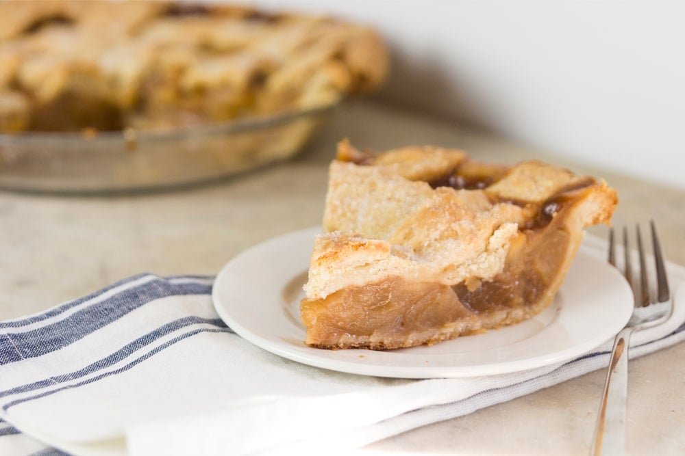 Slice of caramel apple pie sitting on white plate with rest of pie in background all on marble surface with napkin underneath plate