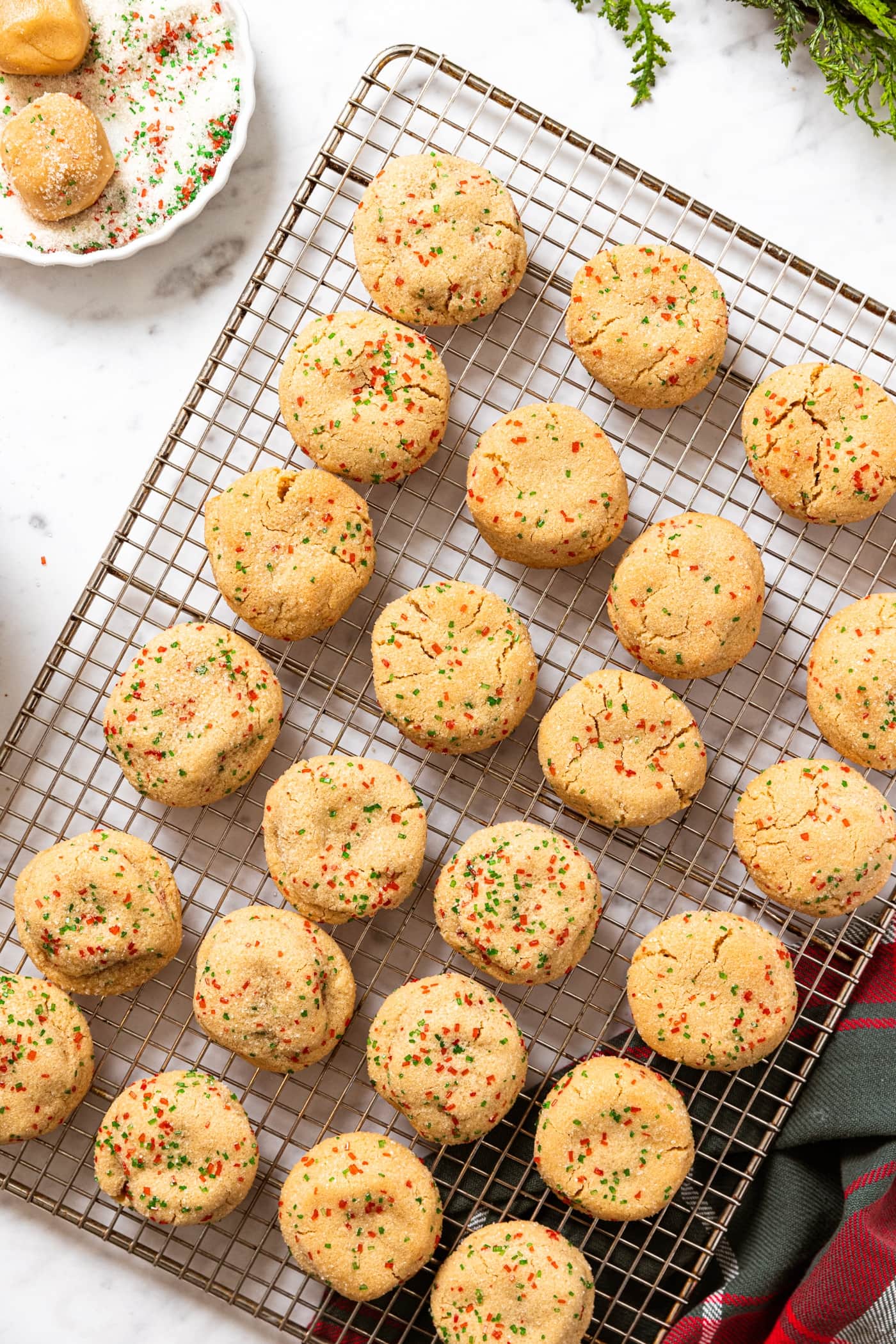 Wire cooling rack filled with light yellow colored cookies dotted with green and red sprinkles all on marble surface with a container filled with sanding sugar with two unbaked cookie dough balls.