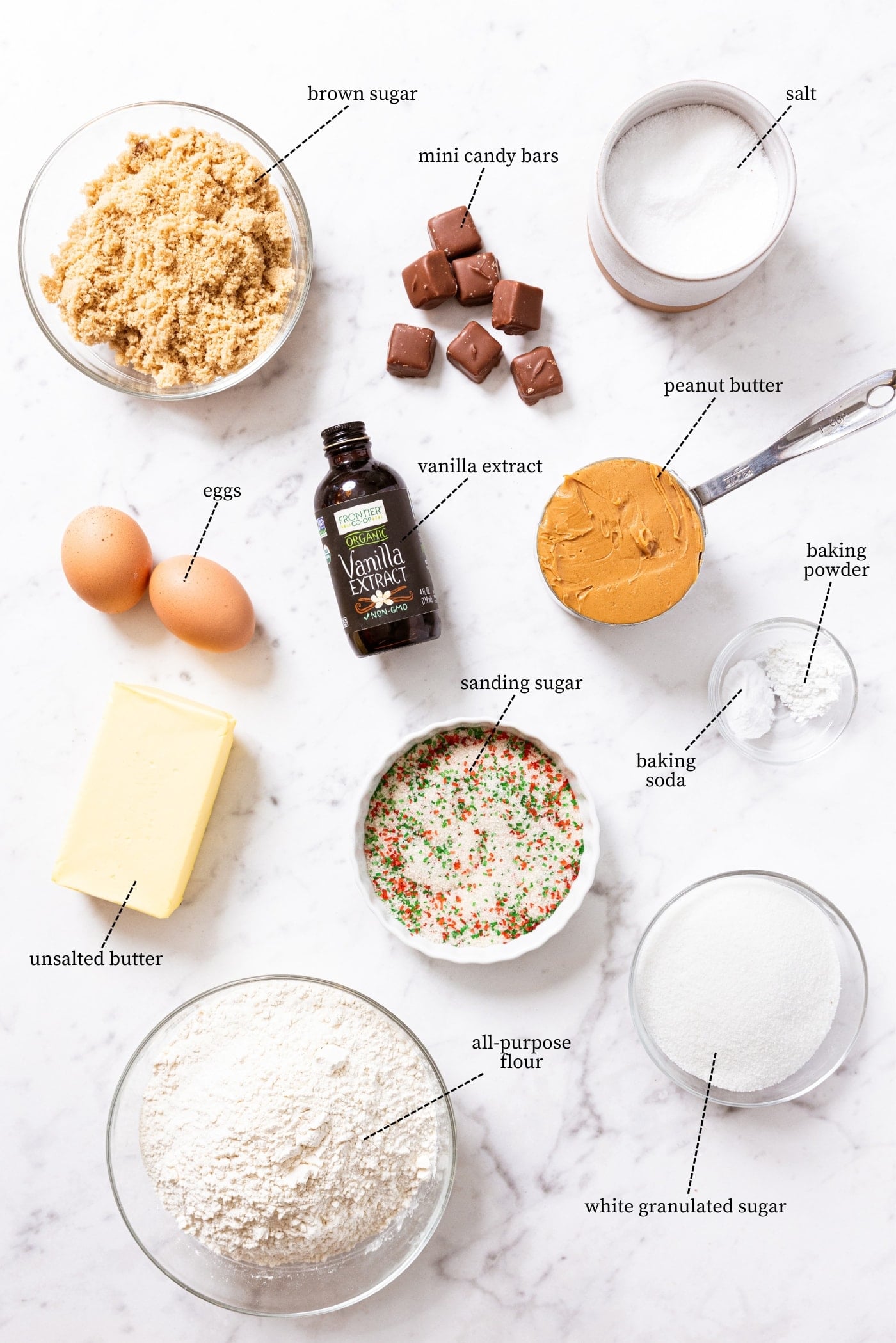 White marble surface with all ingredients needed to make candy bar cookies including mini candy bars, brown sugar, peanut butter, sugar, butter, flour, and more.