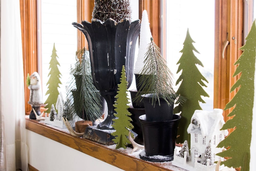 Bow window filled with urns adorned with Christmas decor including cutout green and white trees with fake snow sprinkled around.