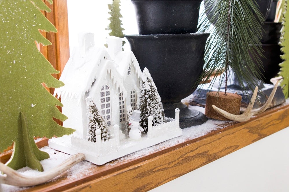 Bow window with urns and white Christmas house covered in snow with green cutout Christmas trees.