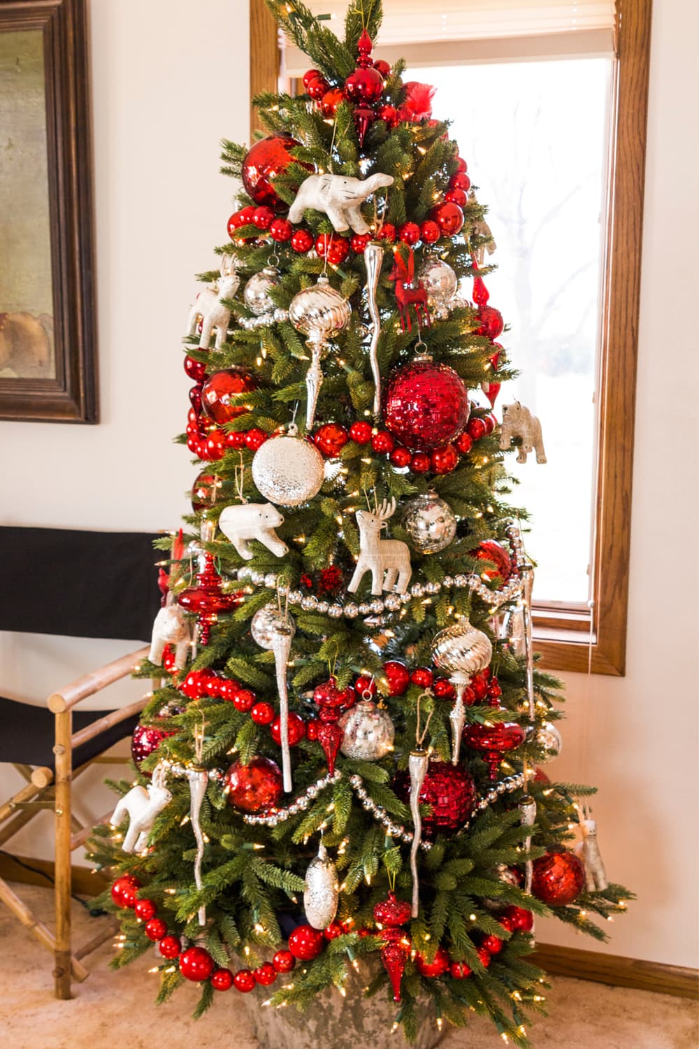 Red and white tree with garland and deer ornaments with chair to the side in front of window.