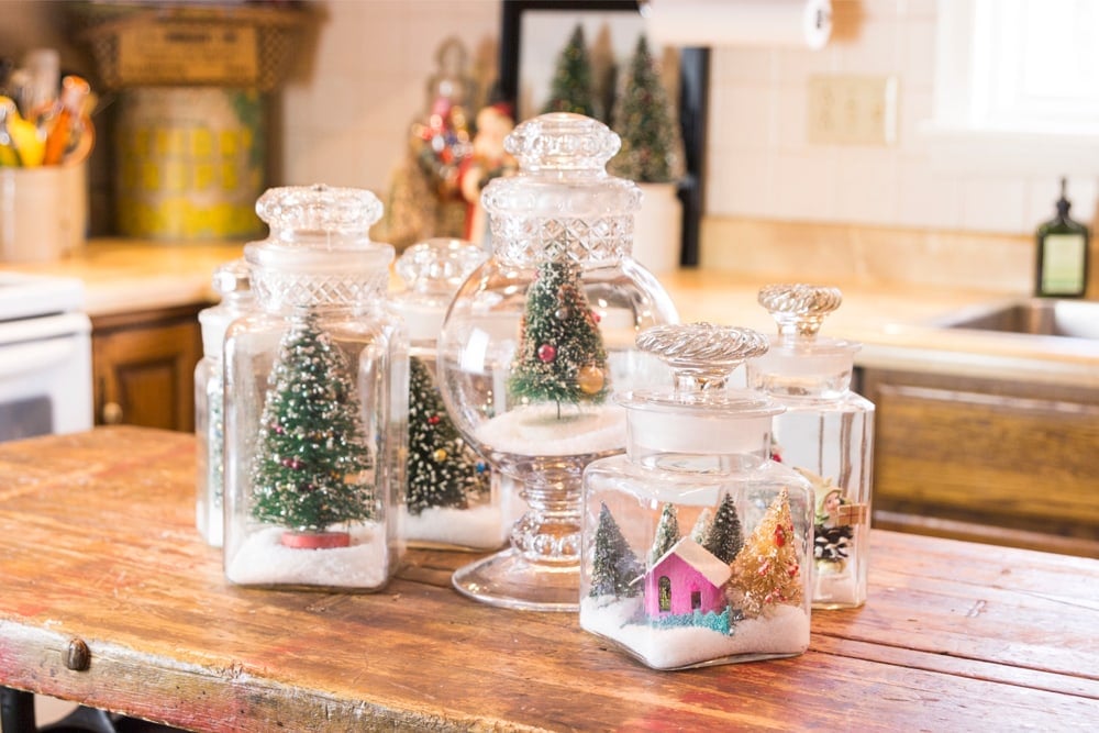 Glass jars filled with fake snow and bottle brush trees and little Putz houses all on wooden kitchen island.