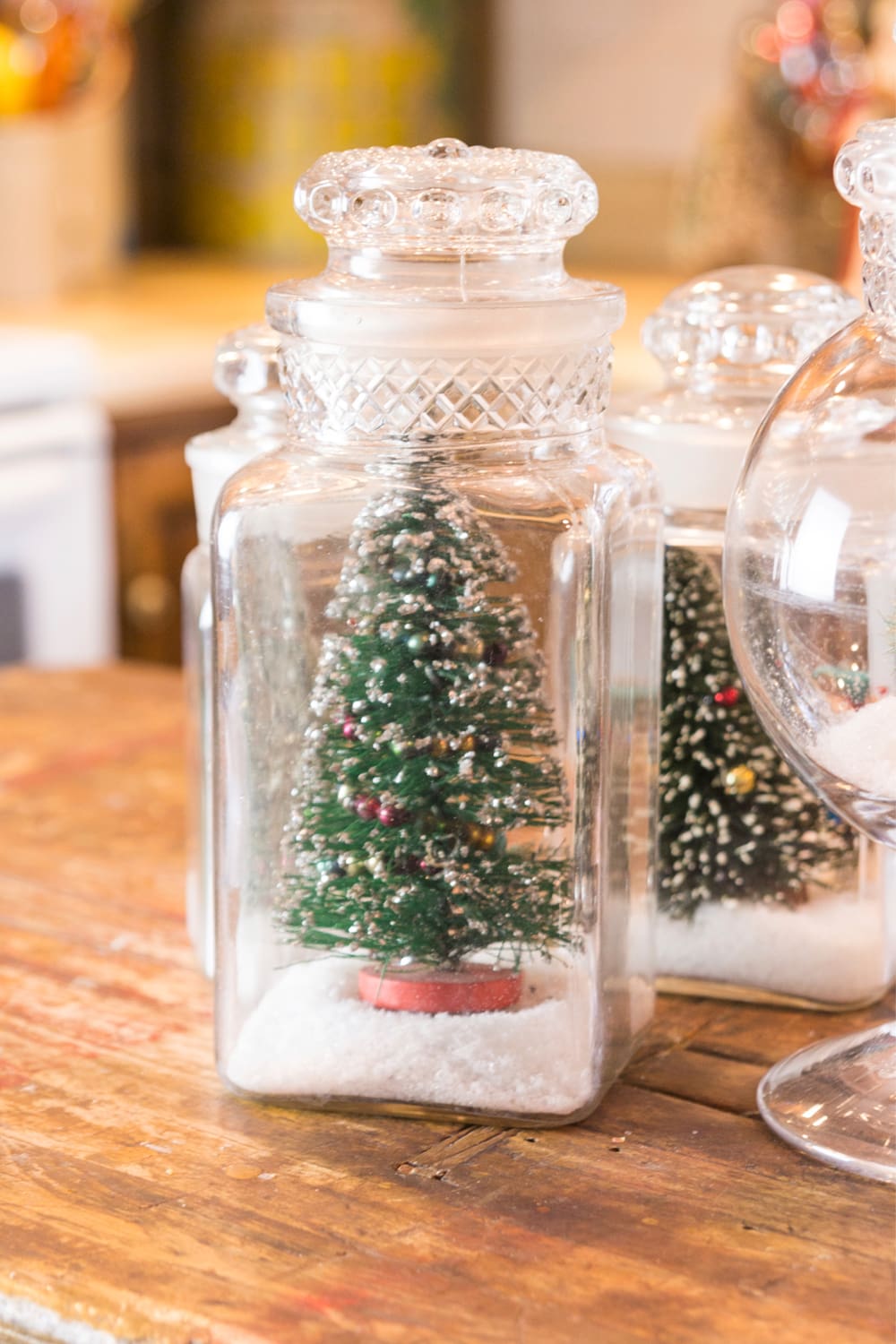 Glass jar filled with fake snow with bottle brush trees resting inside all on wooden island in kitchen.
