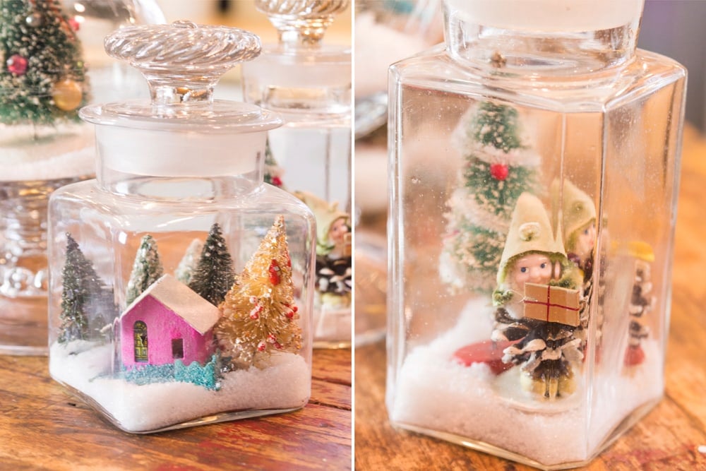 Two glass jars filled with Christmas ornaments resting on fake snow including elf and Putz house.