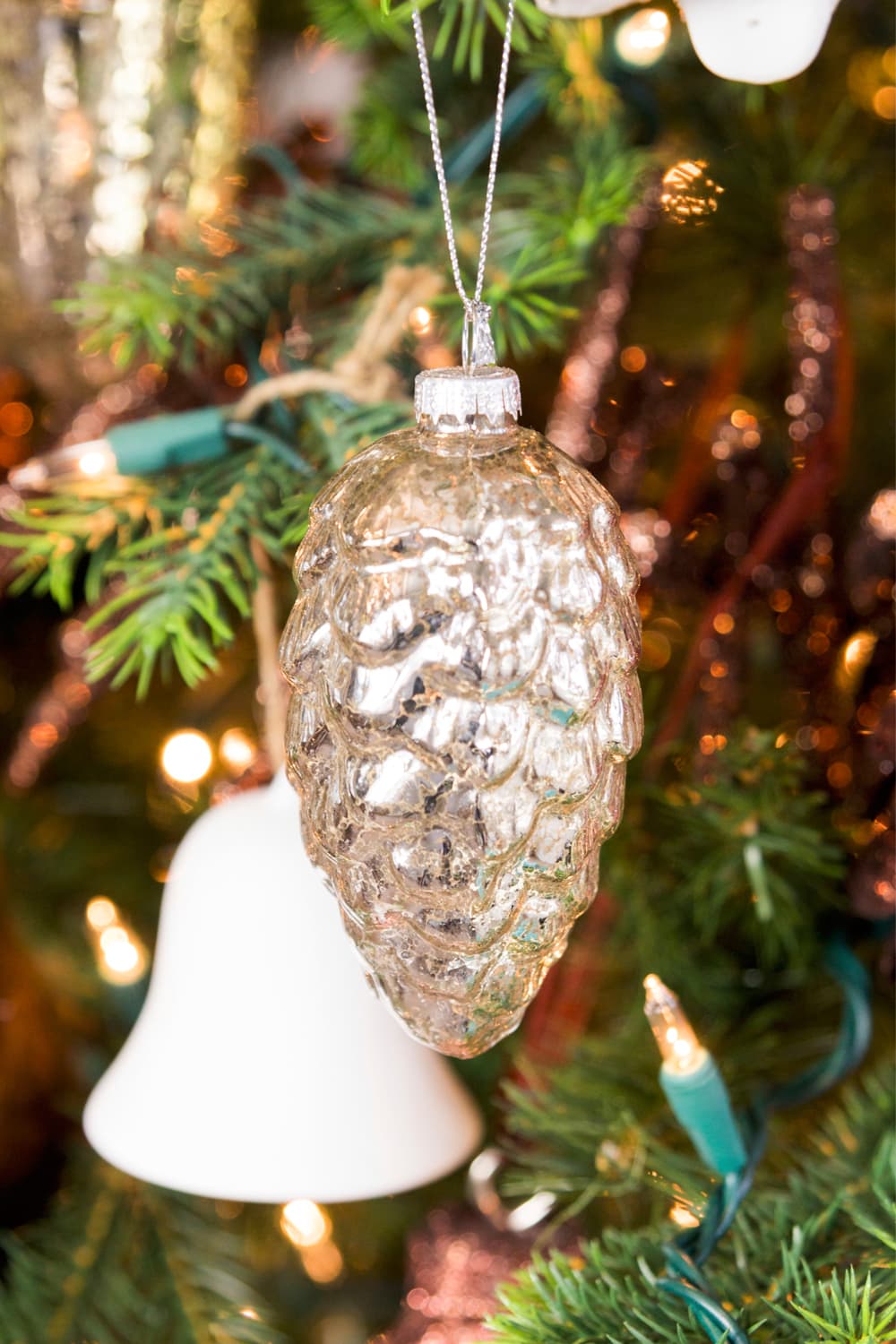 Glass silver mushroom ornament hanging from a bough of a Christmas tree with white bell in the background.