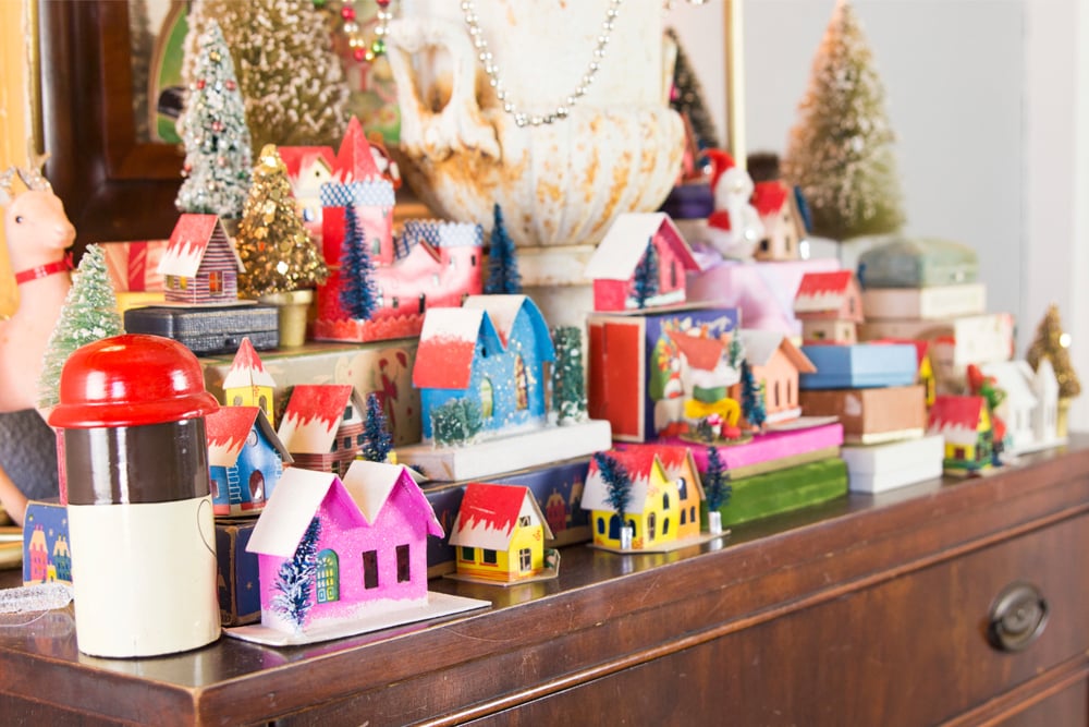 Brown wooden table with lots of holiday and Christmas boxes and Putz houses all over with mirror in background.