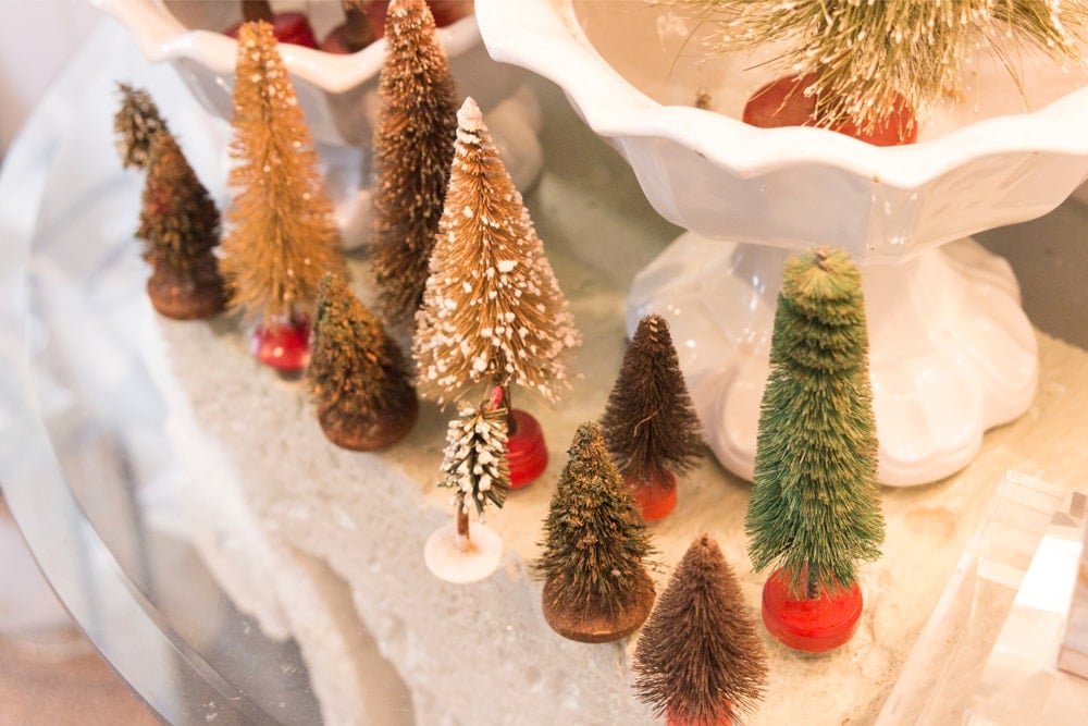Glass table filled with small bottle brush Christmas tree ornaments with white bowl behind.