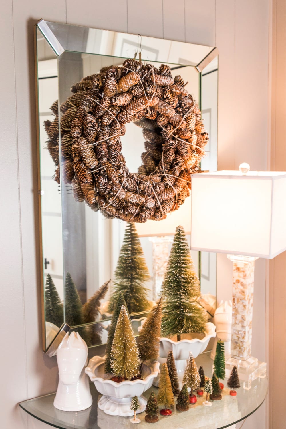 Mirror sitting behind a table with a glass top with pinecone wreath and small bottle brush trees in white bowls with lamp to the side.