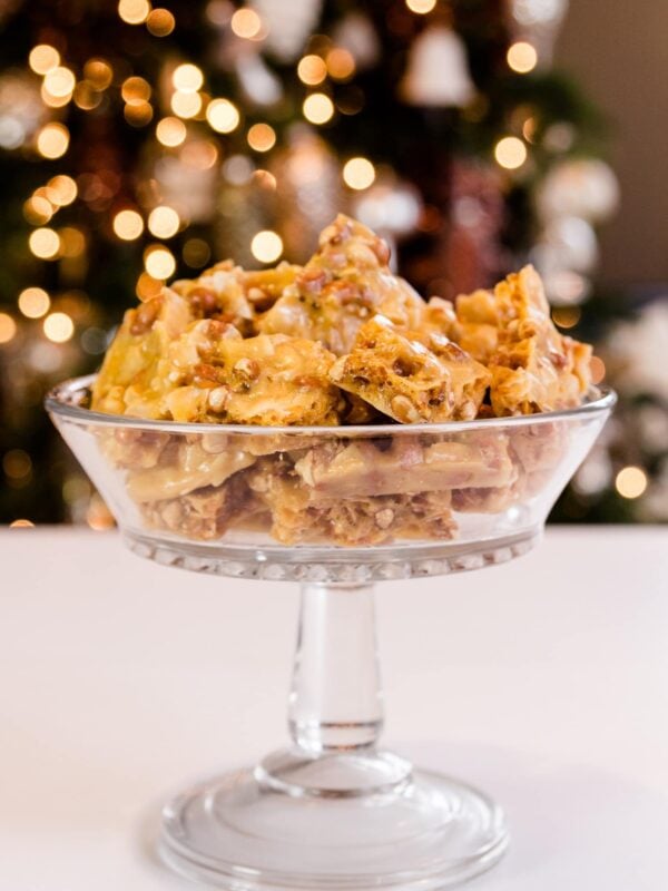 Golden peco candy with peanuts and flakes of coconut piled high in glass bowl on a white surface with Christmas lights in background