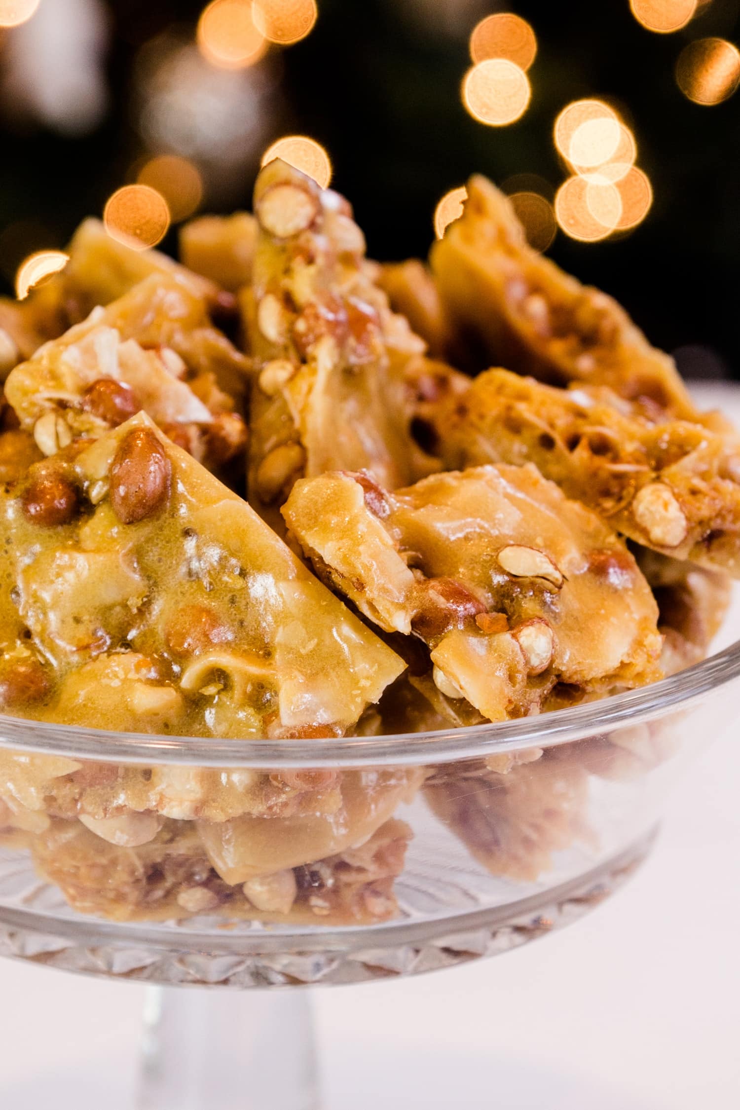 Golden peco candy with peanuts and flakes of coconut piled high in glass bowl on a white surface with Christmas lights in background