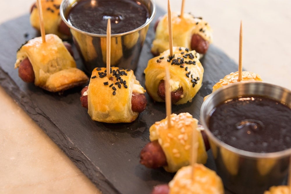 Pigs in a blanket wrapped in browned dough and topped with sesame seeds sitting on slate serving platter with barbecue sauce in steel container all on marble surface