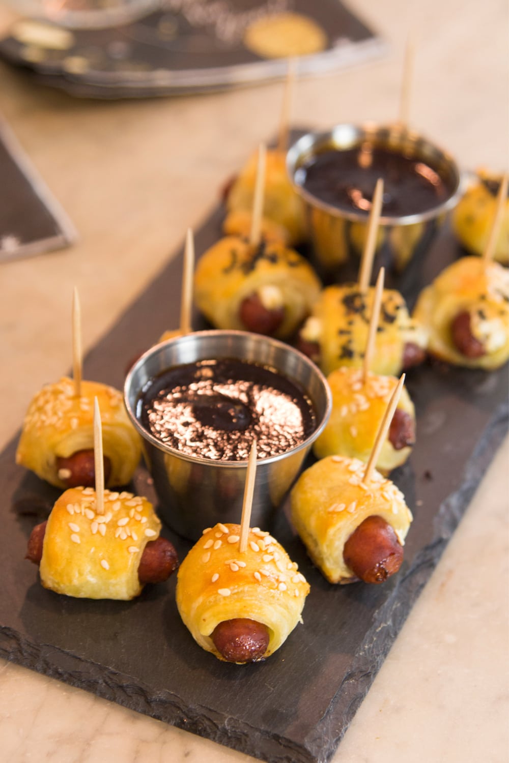 Top down view of pigs in a blanket wrapped in dough and topped with sesame seeds with homemade barbecue sauce in steel container all on slate serving platter set on marble surface