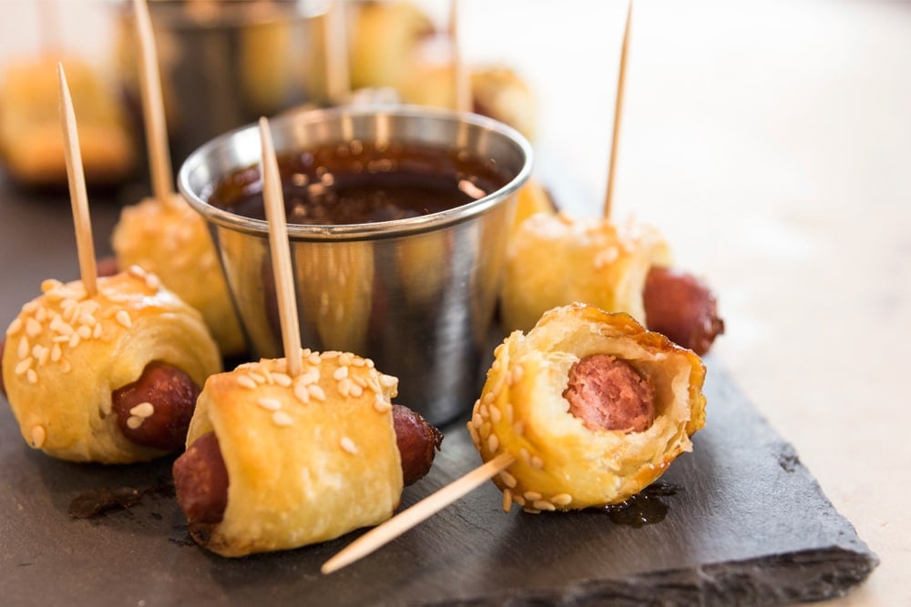 Little smoky hot dogs wrapped in dough with a bite missing with steel container in background holding barbecue sauce all on slate surface