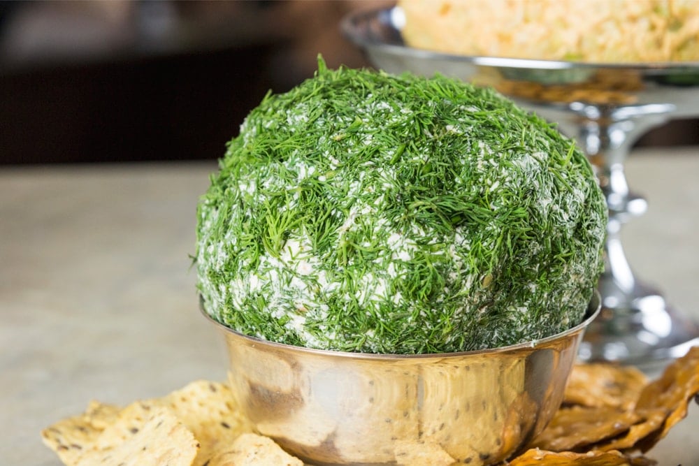 Large cheese ball covered in chopped dill sitting in silver platter with crackers around base