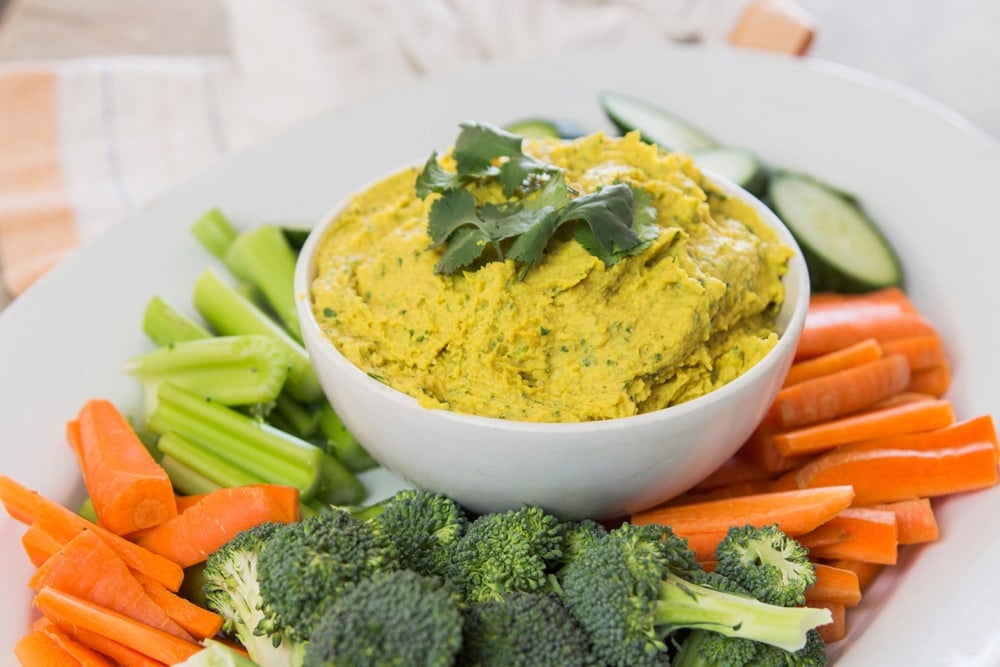 White serving platter filled with sliced vegetables with white bowl filled with curried white bean hummus on top
