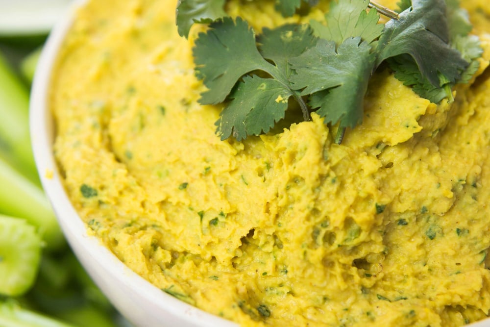 Close up view of yellow colored hummus made from white beans with pieces of cilantro on top with celery in background