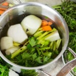 Large steel pot sitting on marble surface filled with greens, onions, carrots, and peppercorns along with lots of water.