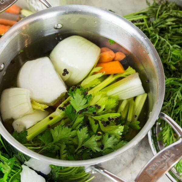 Large steel pot sitting on marble surface filled with greens, onions, carrots, and peppercorns along with lots of water.