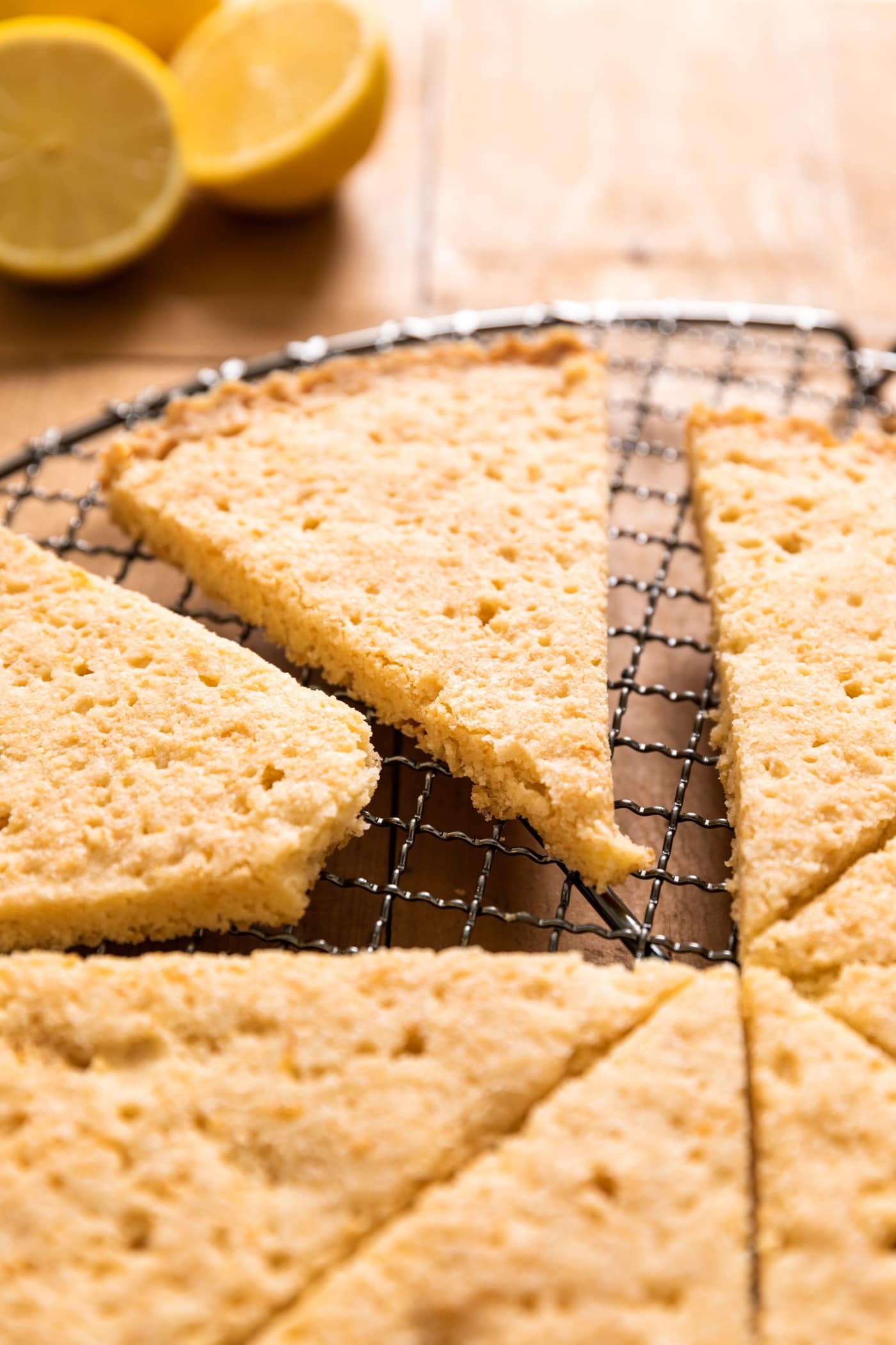 Triangle of golden colored lemon shortbread cookie sitting with other cookies on metal cooling rack with extra lemons in background