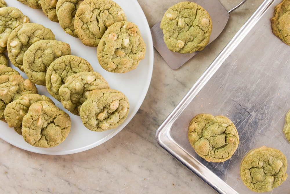 Top down view of white circular serving platter with matcha green tea cookies with white chocolate pieces in them sitting on top with metal baking sheet holding rest of cookies